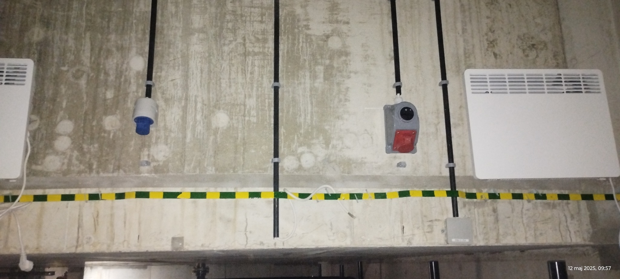 Szara ściana z widocznymi instalacjami elektrycznymi: kable w czarnych rurkach, gniazda przemysłowe, grzejniki elektryczne i taśma ostrzegawcza żółto-zielona.