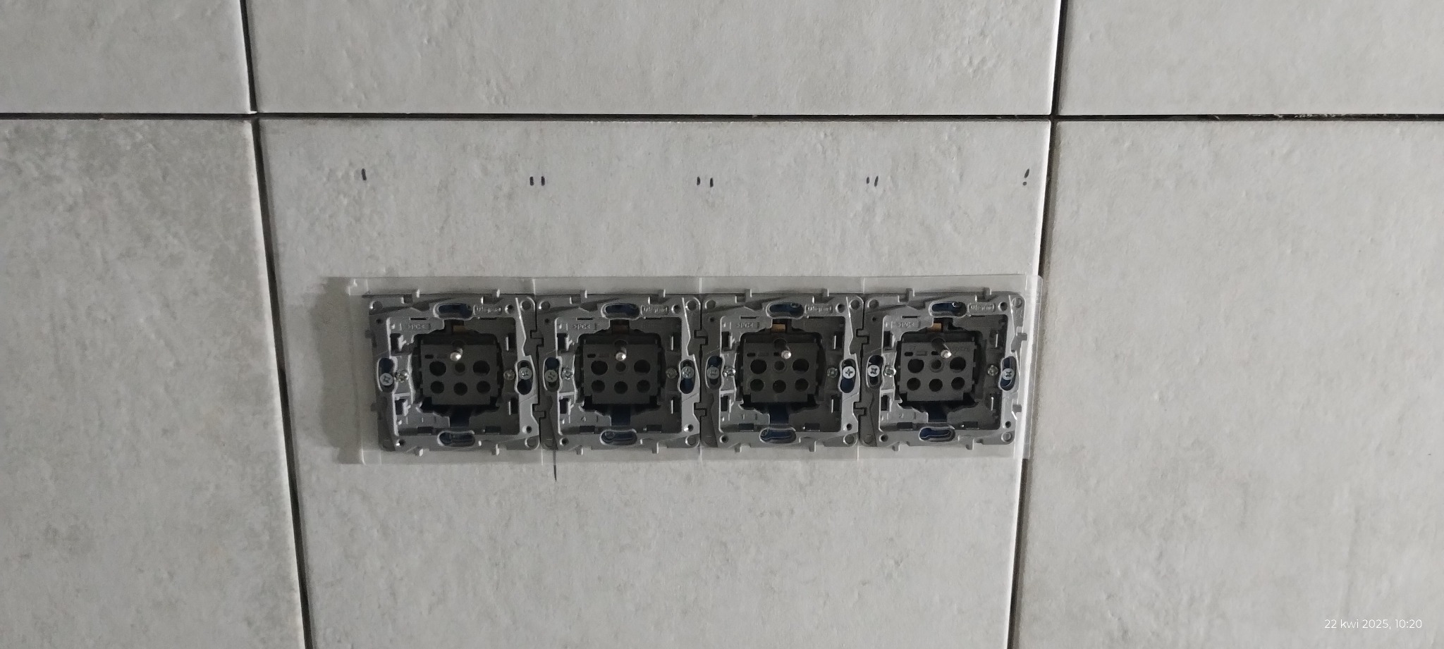 Cztery puszki elektryczne zamontowane w rzędzie na białych, kwadratowych płytkach. Widoczne ślady po ołówku na płytkach.