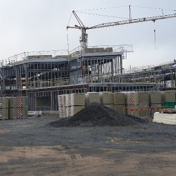 Artur Brzozowski Stombyggen AB - Budowa szkoły i przedszkola w trakcie realizacji: stalowa konstrukcja, materiały izolacyjne, żuraw budowlany na tle pochmurnego nieba. Widok ogólny placu budowy.