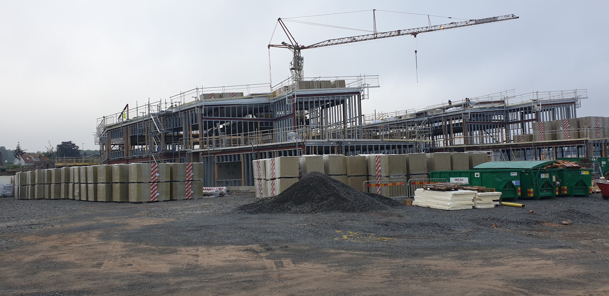 Budowa szkoły i przedszkola w trakcie realizacji: stalowa konstrukcja, materiały izolacyjne, żuraw budowlany na tle pochmurnego nieba. Widok ogólny placu budowy.