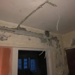 Białe kable elektryczne w bruzdach na ścianie i suficie w trakcie montażu. Widoczne ślady po kuciu w ścianie, surowy stan wykończenia wnętrza.