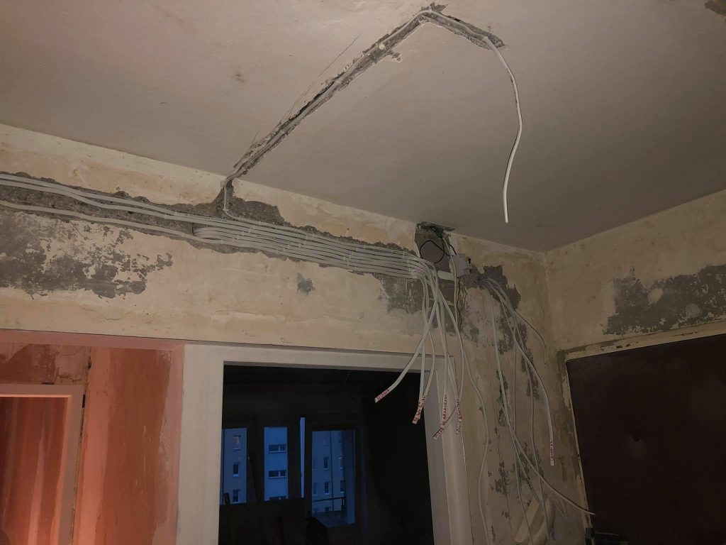 Białe kable elektryczne w bruzdach na ścianie i suficie w trakcie montażu. Widoczne ślady po kuciu w ścianie, surowy stan wykończenia wnętrza.