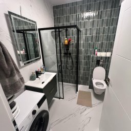 Usługi Remontowo Wykończeniowe BRUN-BUD Karol Kaliniak - Nowoczesna łazienka po remoncie: kabina prysznicowa typu walk-in z czarnymi ramami, umywalka z szafką, pralka i toaleta. Płytki imitujące marmur na podłodze i ścianach.