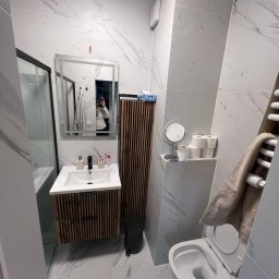 Usługi Remontowo Wykończeniowe BRUN-BUD Karol Kaliniak - Nowoczesna łazienka z marmurowymi płytkami, drewnianą szafką pod umywalką i kabiną prysznicową z czarnymi ramami. W lustrze odbija się osoba robiąca zdjęcie.