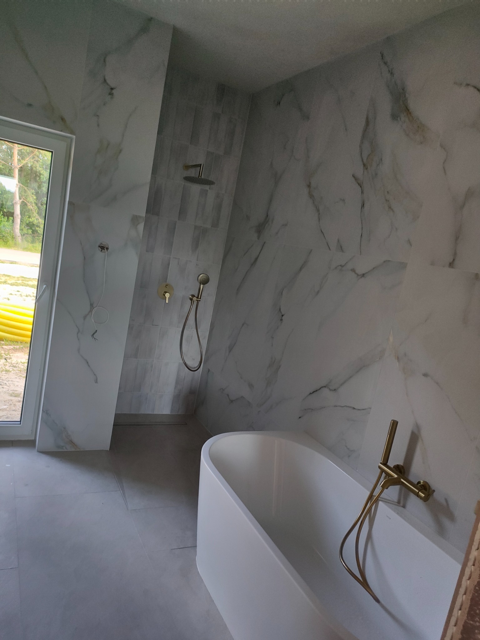 Nowoczesna łazienka z wanną, marmurowymi płytkami i złotą armaturą. Widok na okno z widokiem na zieleń. Minimalistyczny design, jasne kolory.