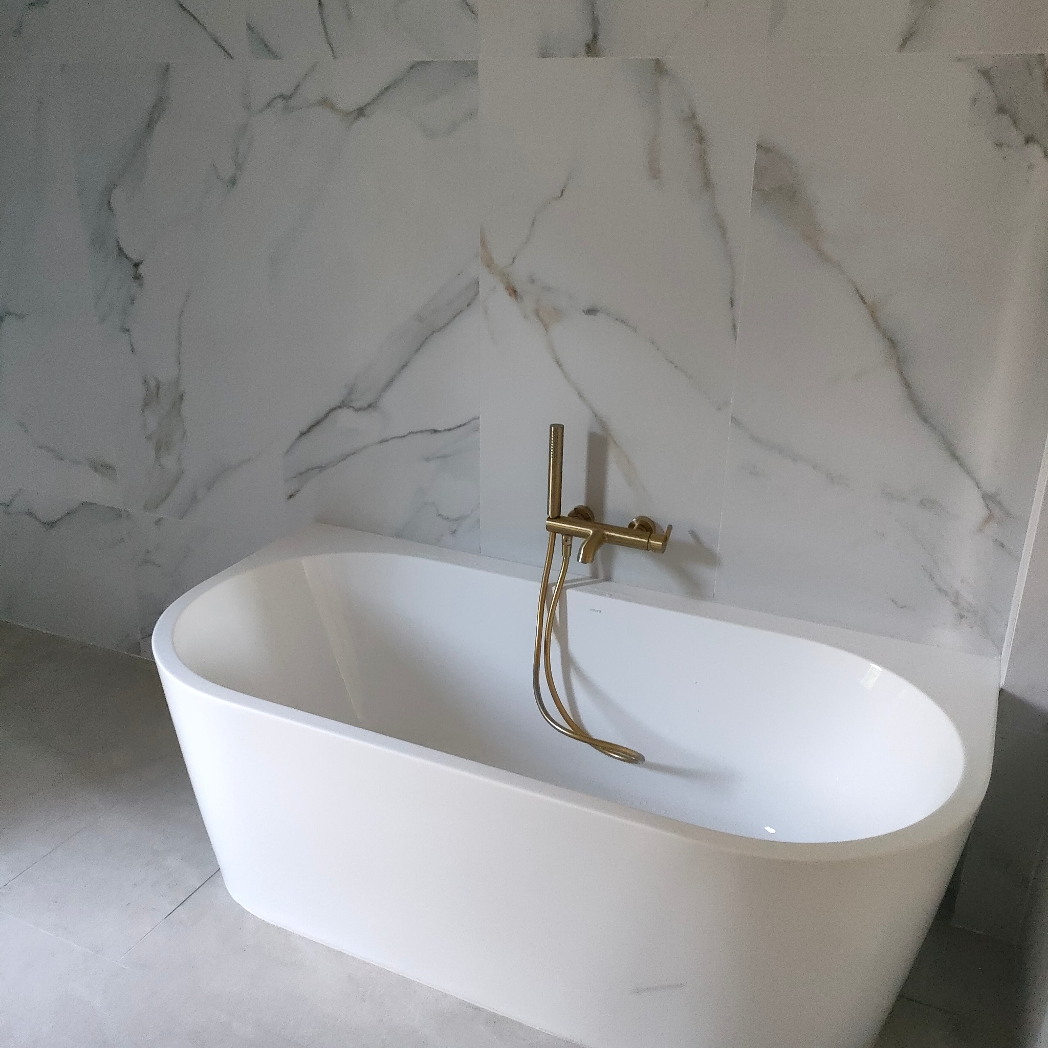 Biała wanna z złotą armaturą na tle ściany z płytek imitujących marmur w łazience w Siennicy Nadolnej. Nowoczesne wykończenie.