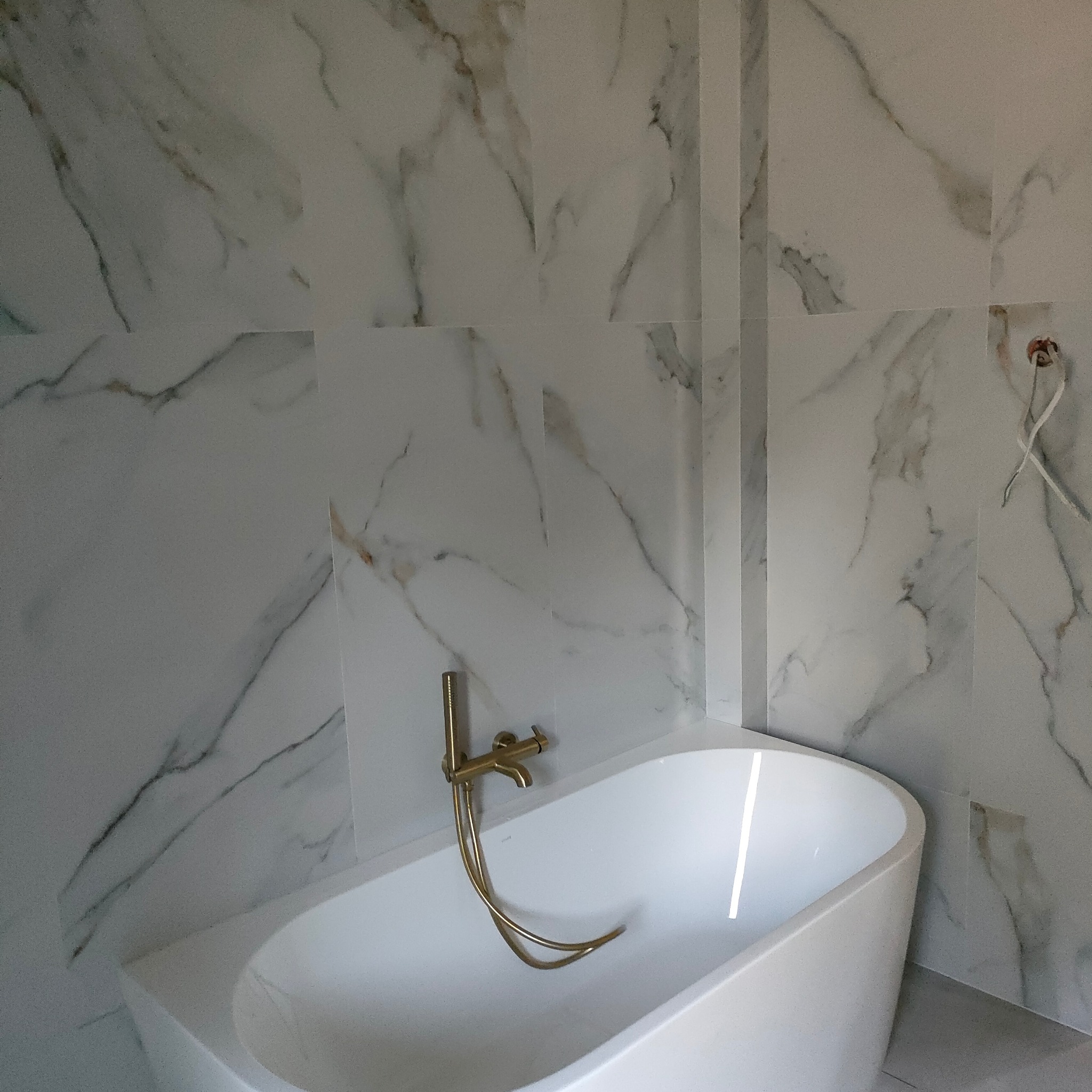 Biała wanna wolnostojąca z złotą armaturą na tle marmurowych płytek. Widoczne niedokończone podłączenie elektryczne. Minimalistyczny design łazienki.