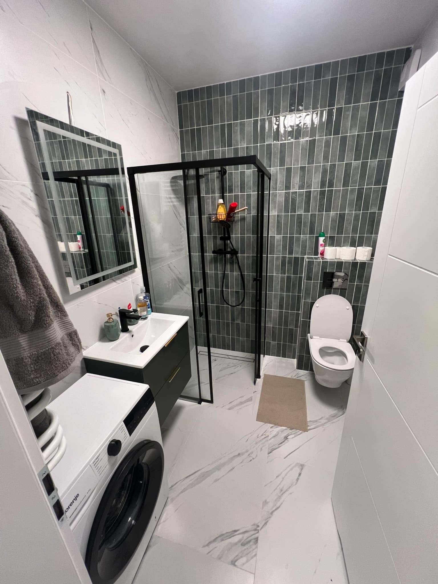 Nowoczesna łazienka po remoncie: kabina prysznicowa typu walk-in z czarnymi ramami, umywalka z szafką, pralka i toaleta. Płytki imitujące marmur na podłodze i ścianach.