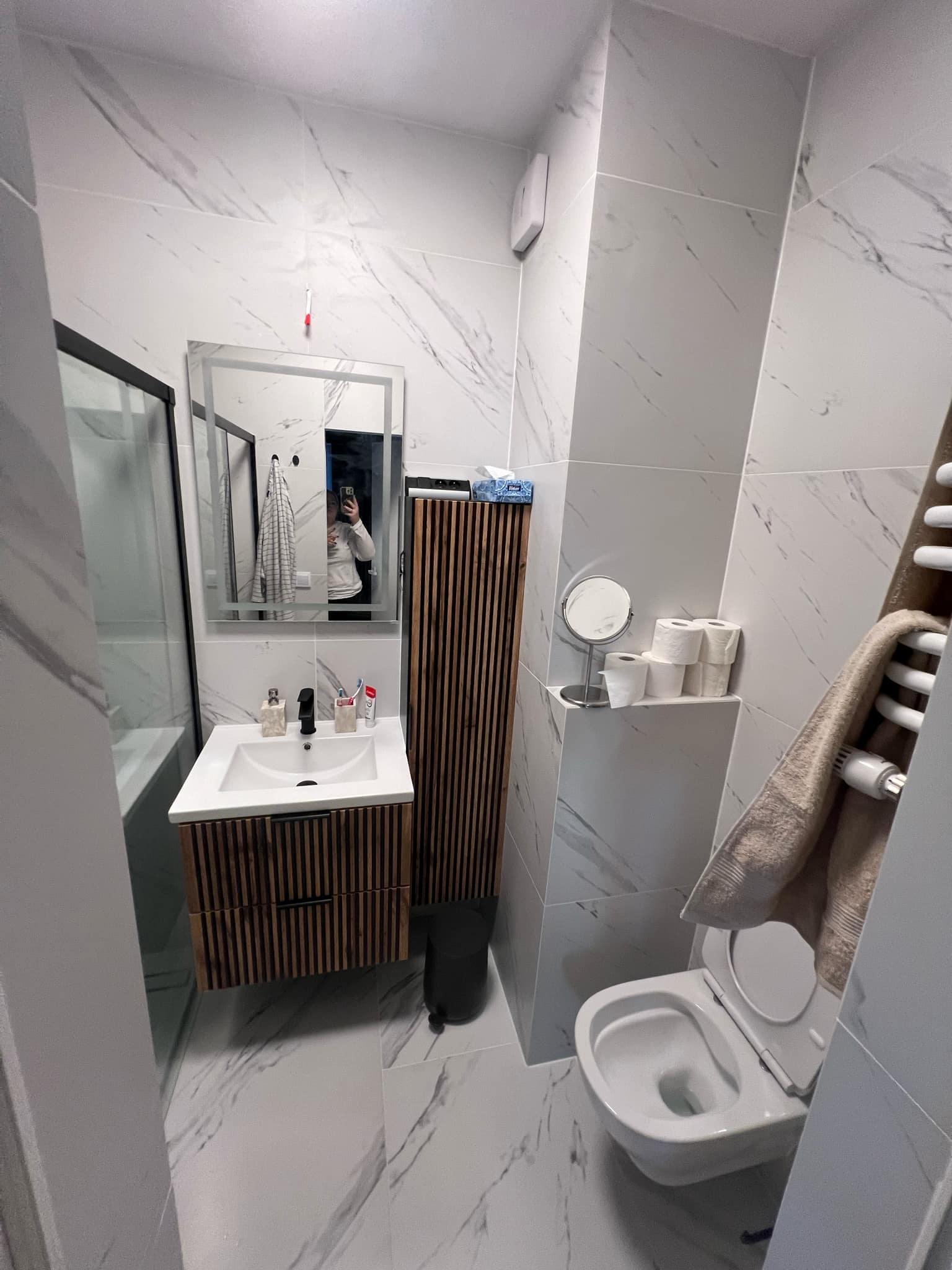 Nowoczesna łazienka z marmurowymi płytkami, drewnianą szafką pod umywalką i kabiną prysznicową z czarnymi ramami. W lustrze odbija się osoba robiąca zdjęcie.