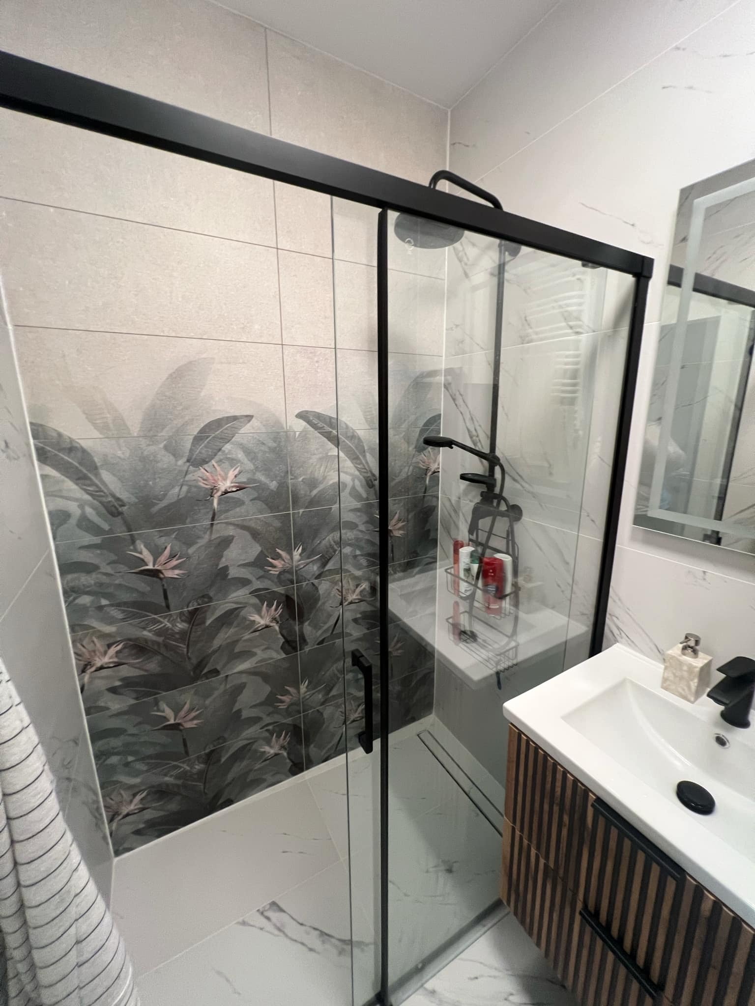 Nowoczesna łazienka z kabiną prysznicową typu walk-in, płytki z motywem roślinnym, umywalka z szafką z lamelami i czarną armaturą. Minimalistyczny design.