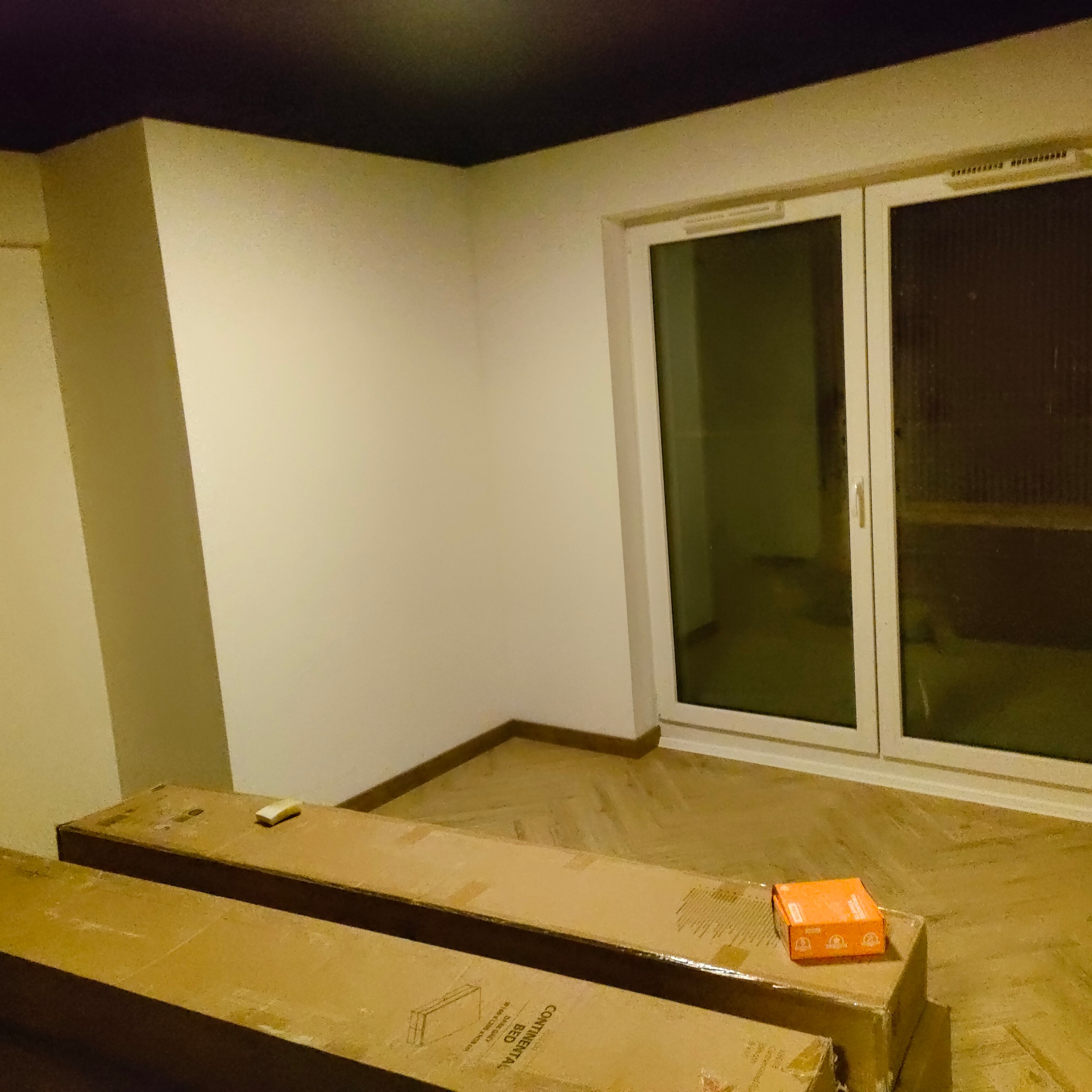 Pokój z parkietem w jodełkę, przygotowany do montażu. Widoczne zapakowane panele podłogowe i pudełko, ściany pomalowane na biało, czarny sufit i okno.