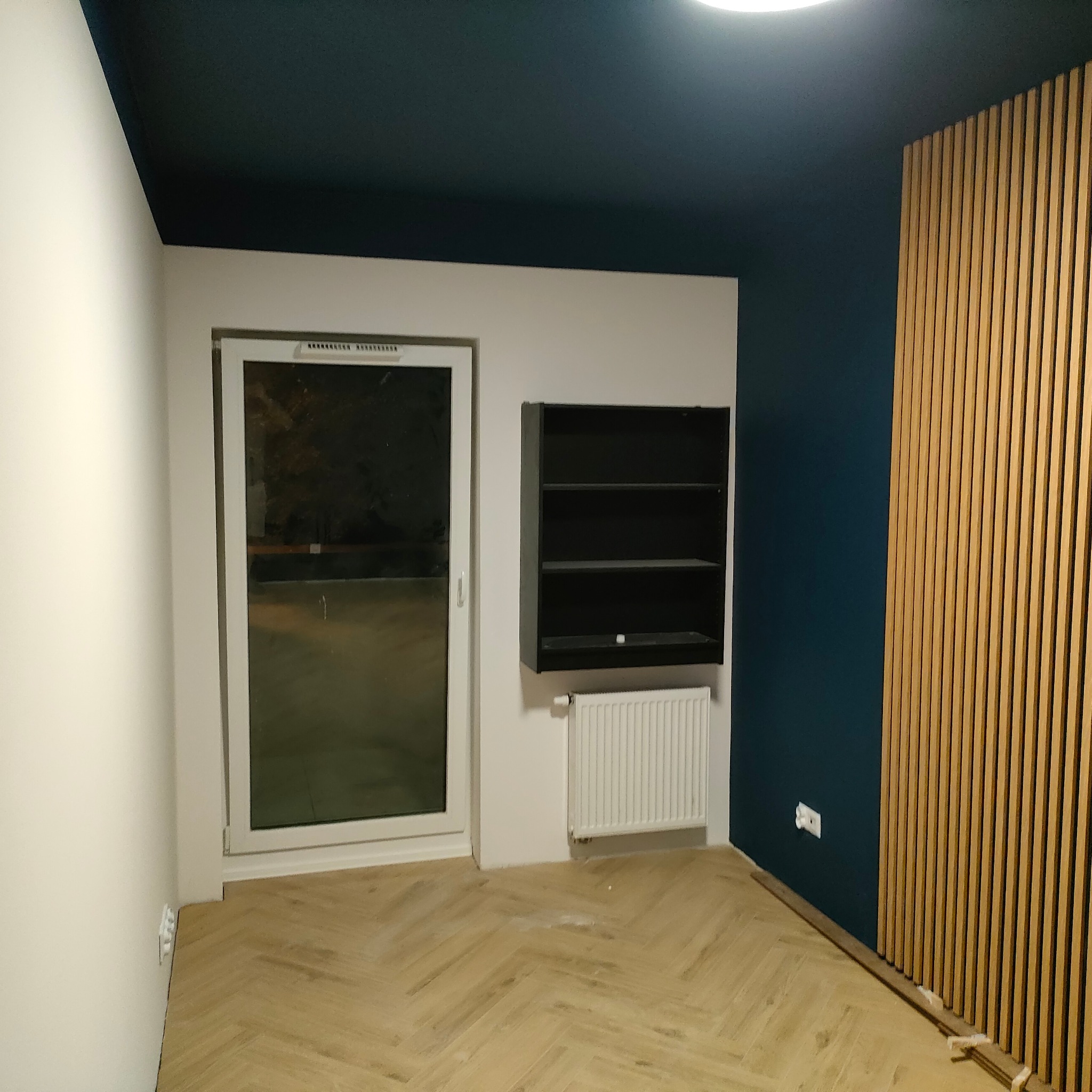 Wnętrze pokoju po remoncie: parkiet w jodełkę, lamela na ścianie, ciemny sufit, białe ściany, okno i czarna półka. Widoczny grzejnik i gniazdko elektryczne.