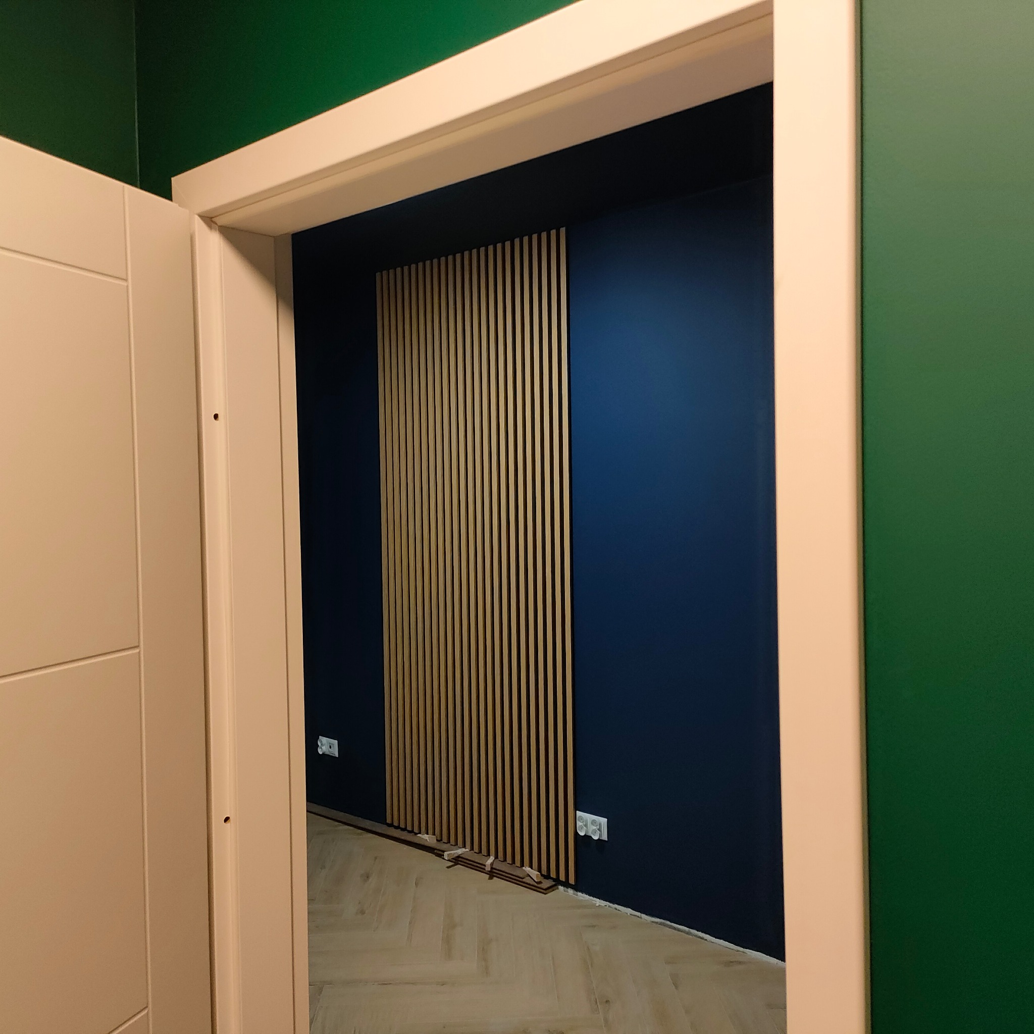 Nowoczesny panel lamelowy na granatowej ścianie, widoczny zza beżowej futryny drzwi. Podłoga ułożona w jodełkę. Kontrastujące kolory ścian: granatowy i butelkowa zieleń.