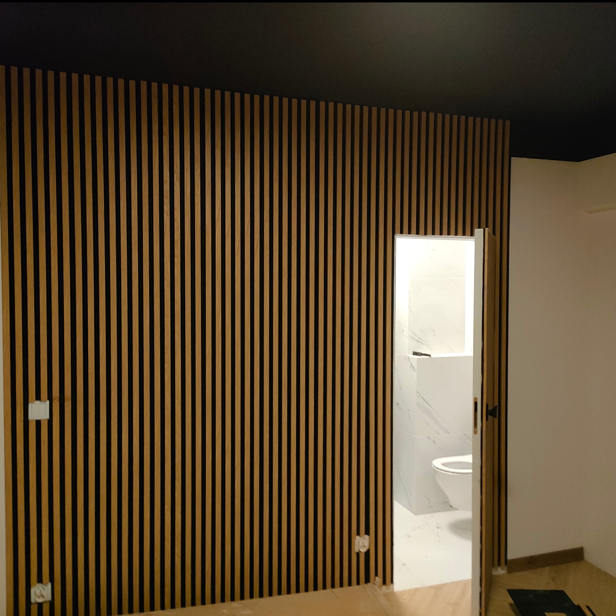 Nowoczesna ściana z drewnianych lameli w kolorze jasnego drewna na czarnym tle, z widocznym wejściem do wykończonej łazienki z białymi płytkami.