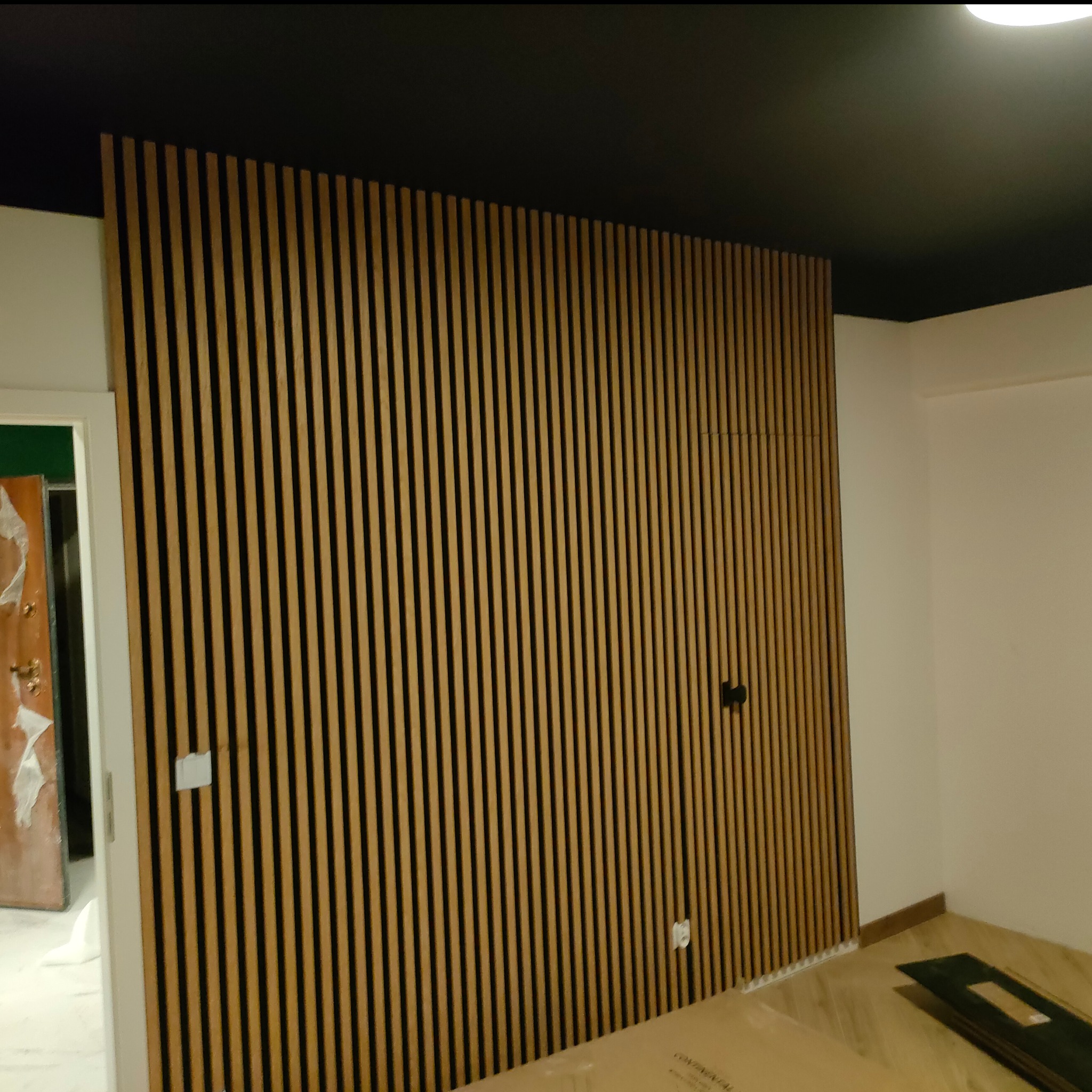 Ściana z drewnianych paneli lamelowych w wykończonym wnętrzu. Widoczne elementy instalacji elektrycznej i czarny sufit. Wykończenie wnętrza w stylu nowoczesnym.