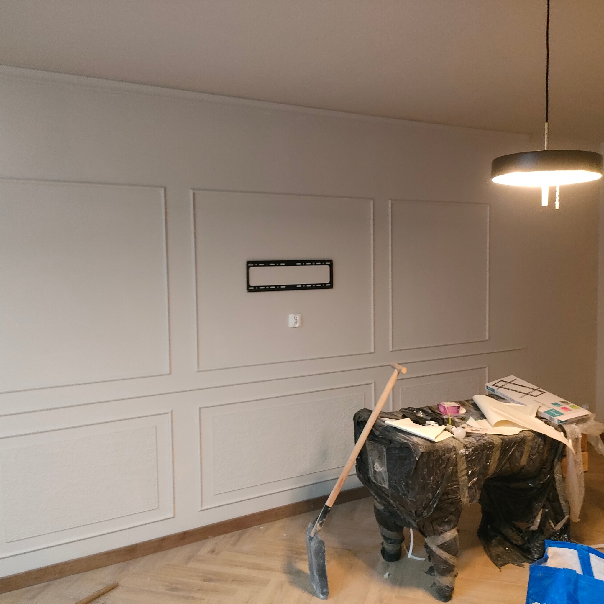 Ściana z dekoracyjnymi panelami i zamontowanym uchwytem TV. Pod ścianą prowizoryczny stół roboczy z narzędziami. Parkiet ułożony w jodełkę.