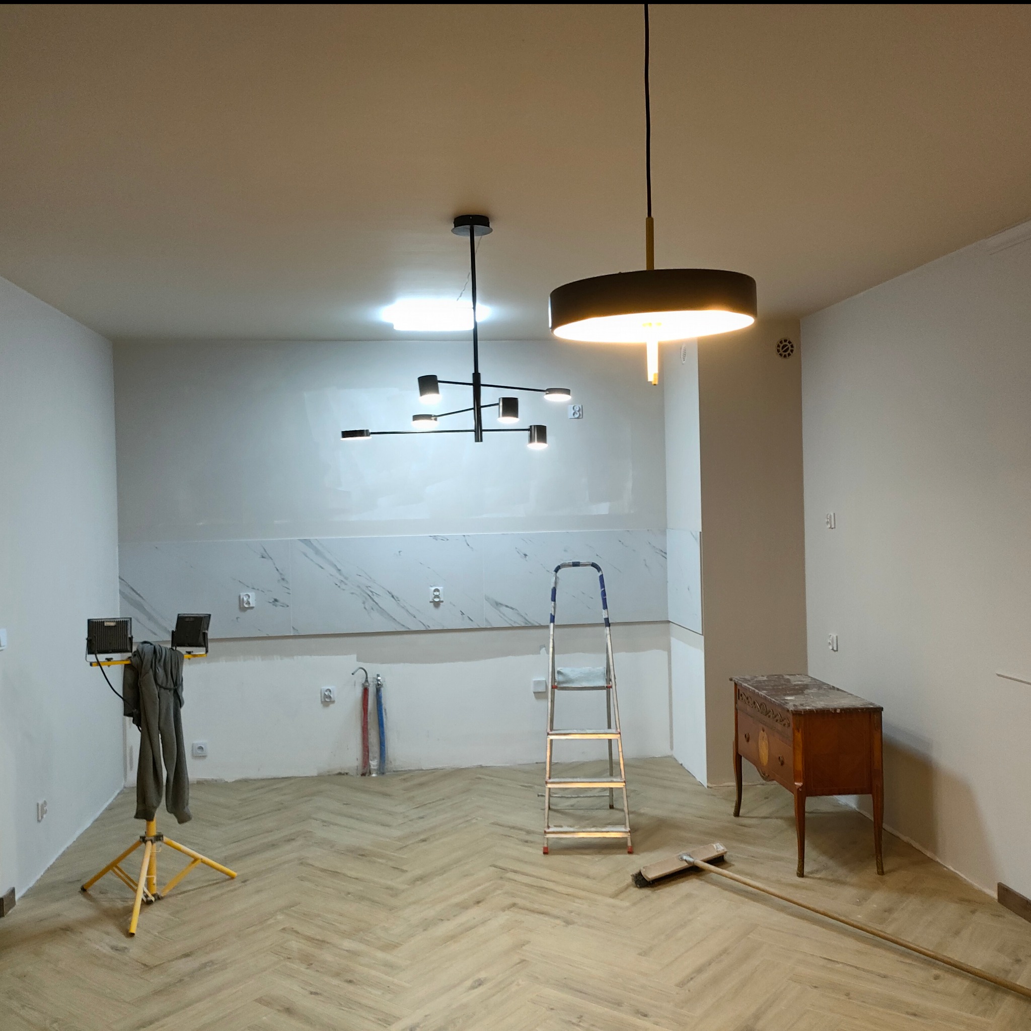 Wnętrze w trakcie prac wykończeniowych: ułożony parkiet w jodełkę, częściowo wykończona ściana z marmurowymi płytkami, drabina i lampy sufitowe.