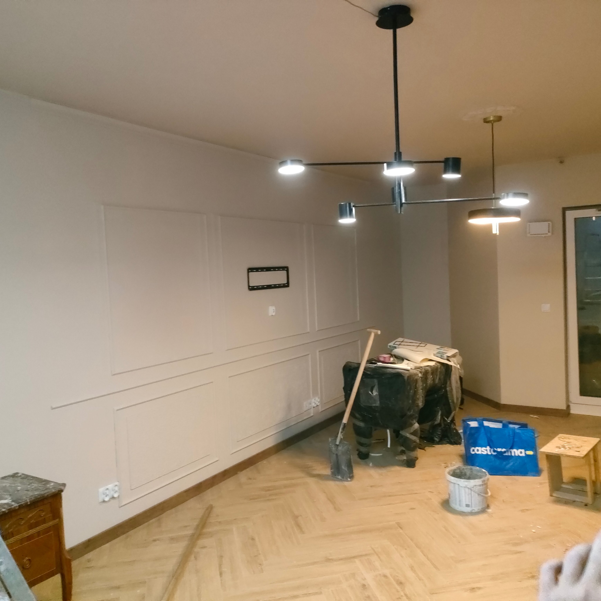 Jasne parkiet ułożony w jodełkę, ściany z dekoracyjną sztukaterią. Widoczne narzędzia i materiały budowlane, nowoczesne oświetlenie sufitowe. Remont wnętrza w trakcie.