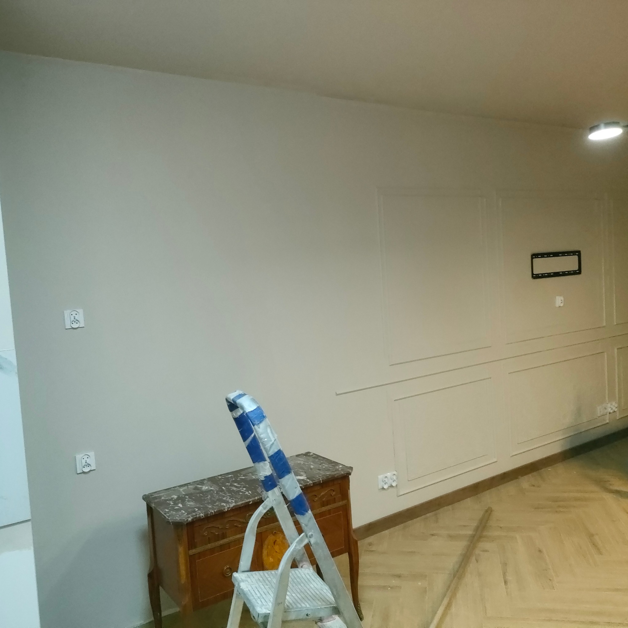 Wnętrze w trakcie prac wykończeniowych: pomalowane ściany, sztukateria, parkiet w jodełkę, komoda, drabina malarska zabezpieczona taśmą. Widoczne gniazdka.