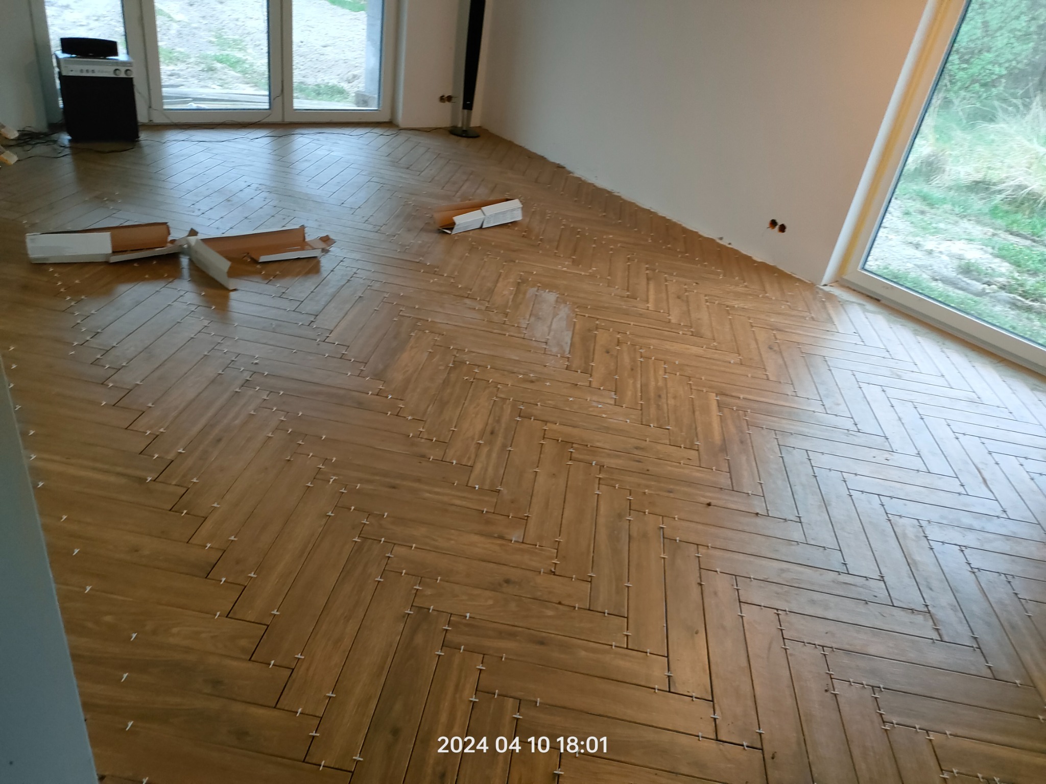 Układanie parkietu w jodełkę z drewnopodobnych płytek. Widoczne krzyżyki dystansowe i puste kartony po płytkach. Jasne wnętrze z dużymi oknami.