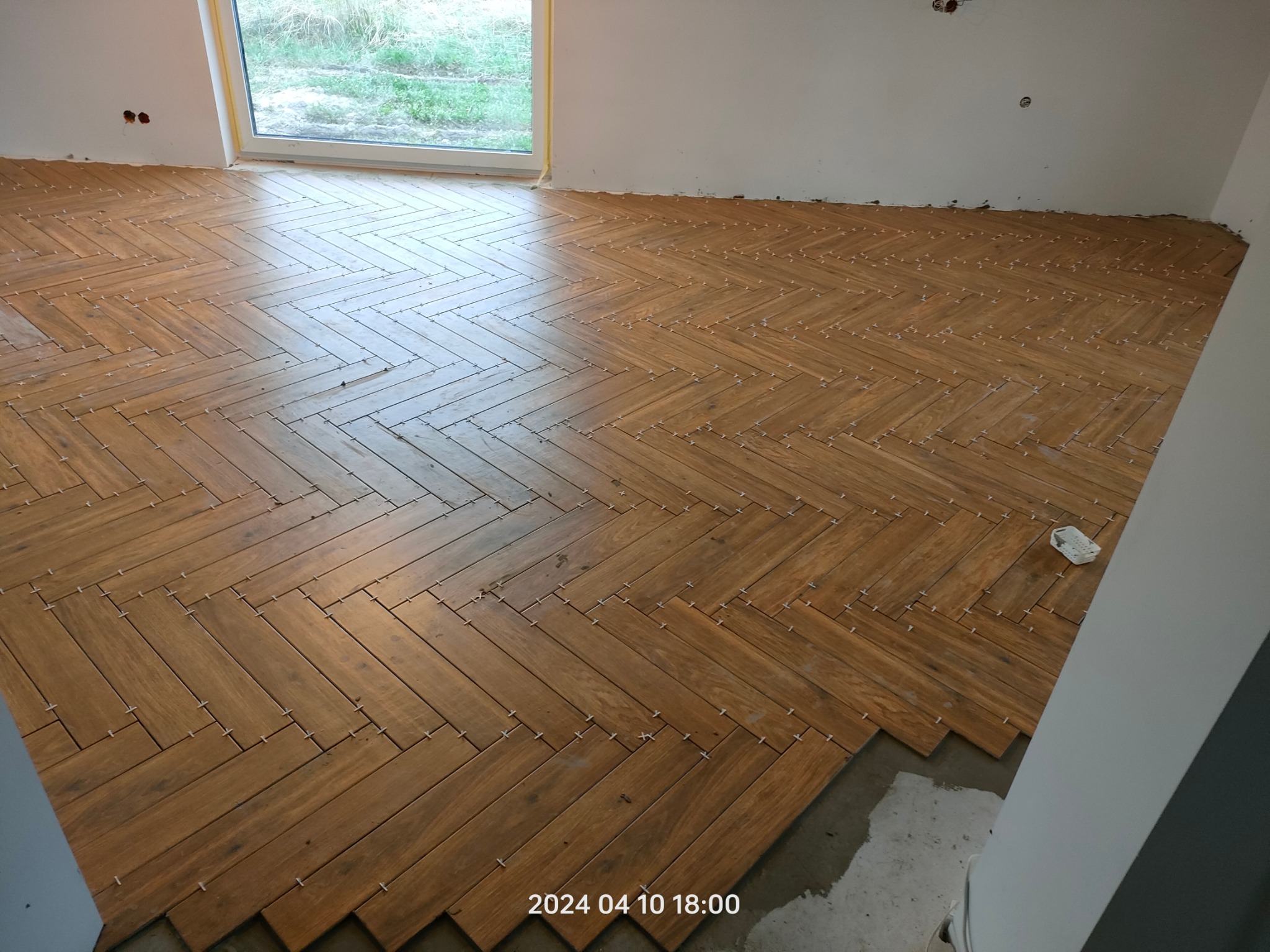 Układanie drewnopodobnych płytek w jodełkę na podłodze w pokoju. Widoczne krzyżyki dystansowe. Częściowo ułożona podłoga, okno w tle, surowy stan.