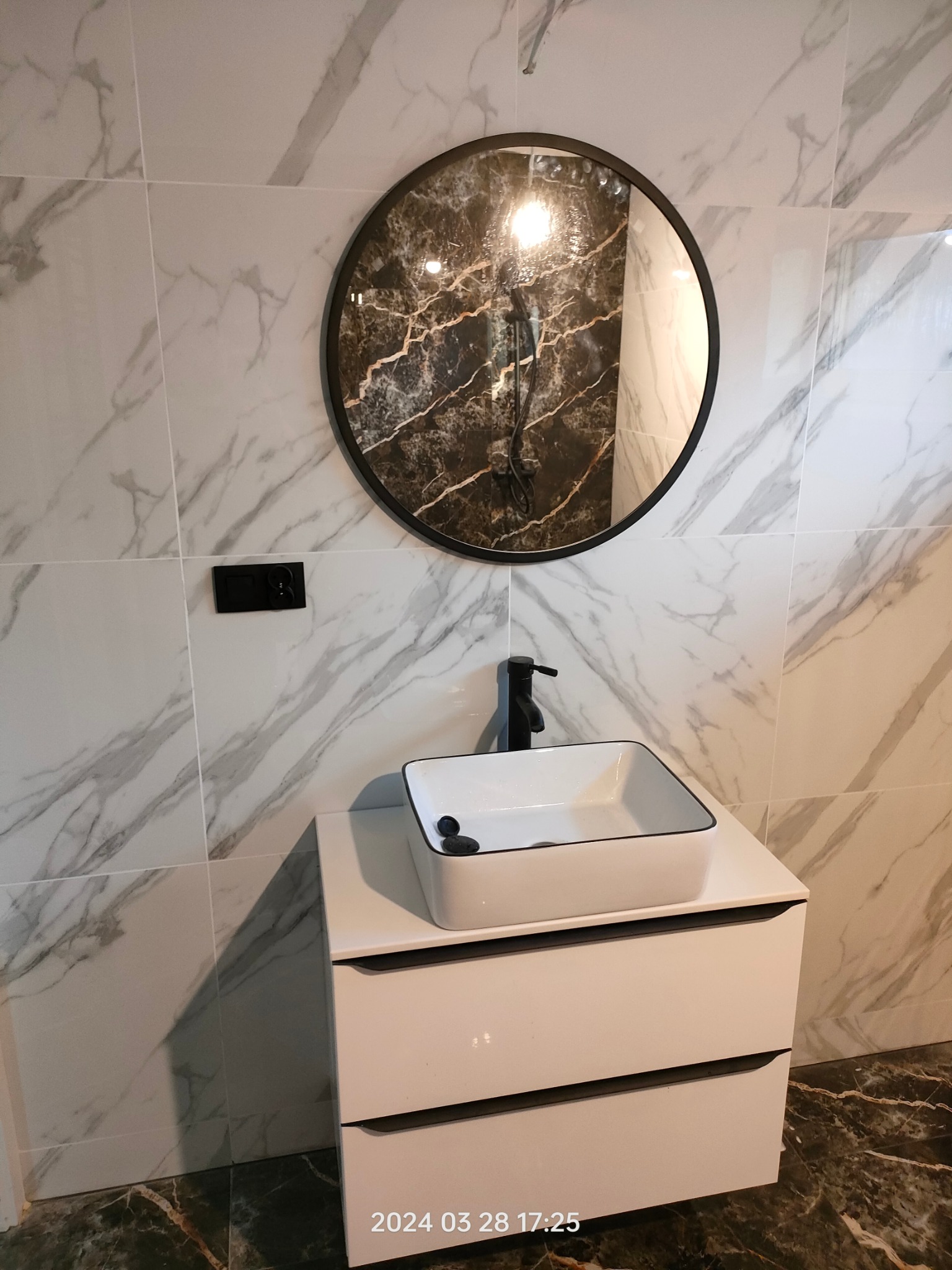 Biała, prostokątna umywalka nablatowa z czarną armaturą na szafce w łazience z marmurowymi płytkami i okrągłym lustrem z ciemnym wzorem.