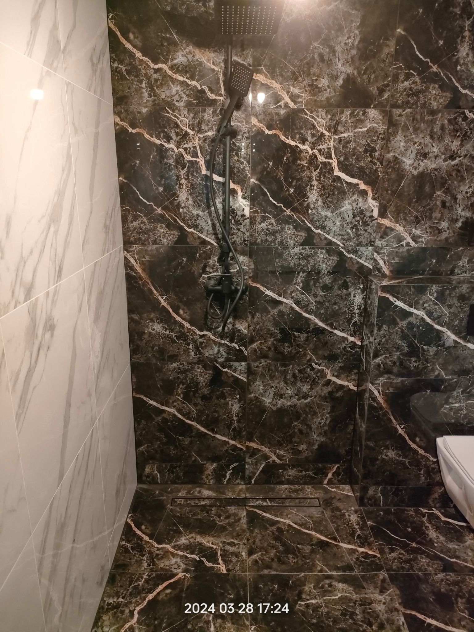 Nowoczesna kabina prysznicowa z czarnymi płytkami imitującymi marmur i czarną armaturą, z odpływem liniowym. Kontrastuje z jasną, marmurową ścianą.