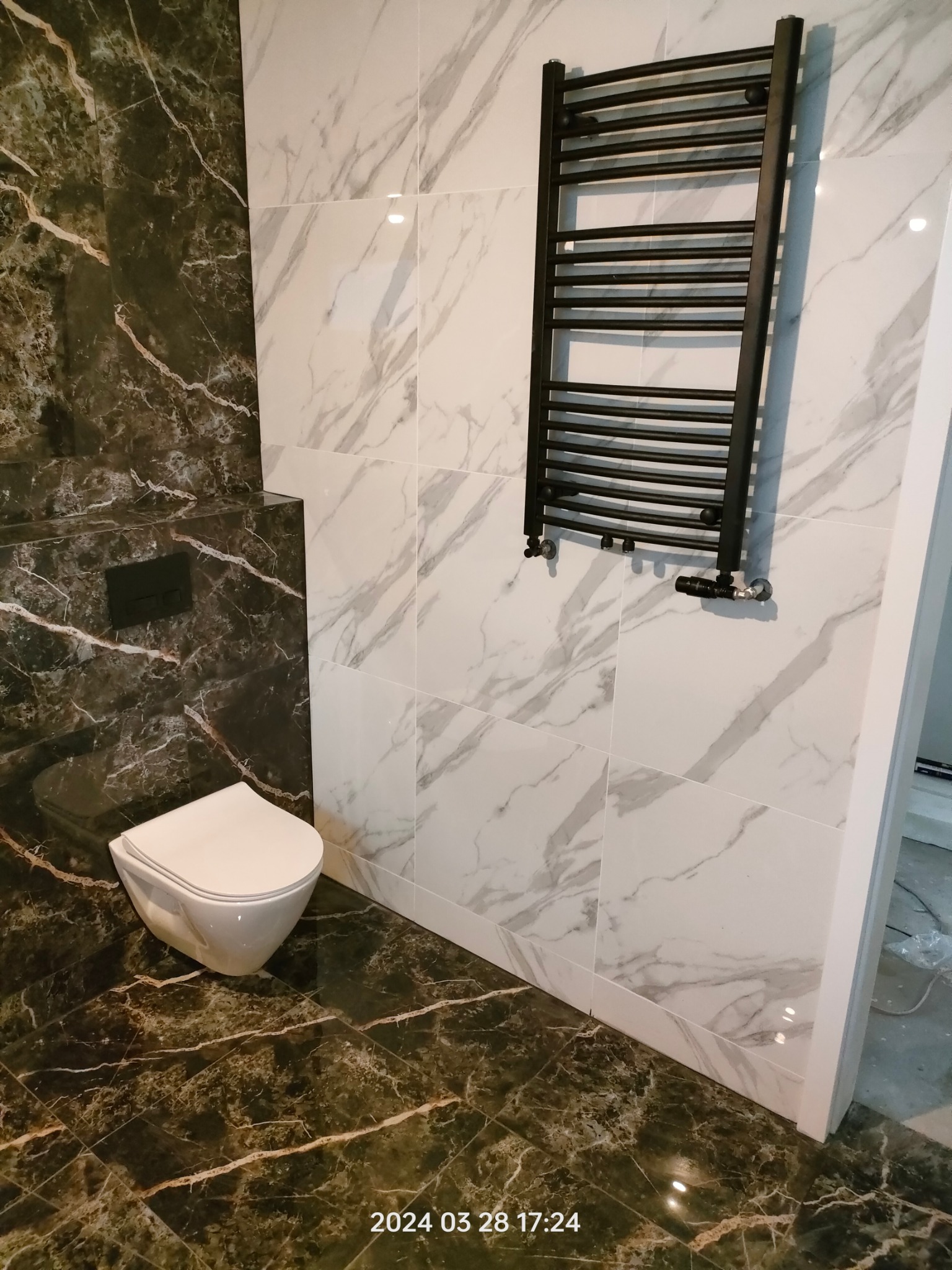 Elegancka łazienka po metamorfozie: marmurowe płytki w odcieniach bieli i czerni, wisząca toaleta oraz designerski, czarny grzejnik drabinkowy.