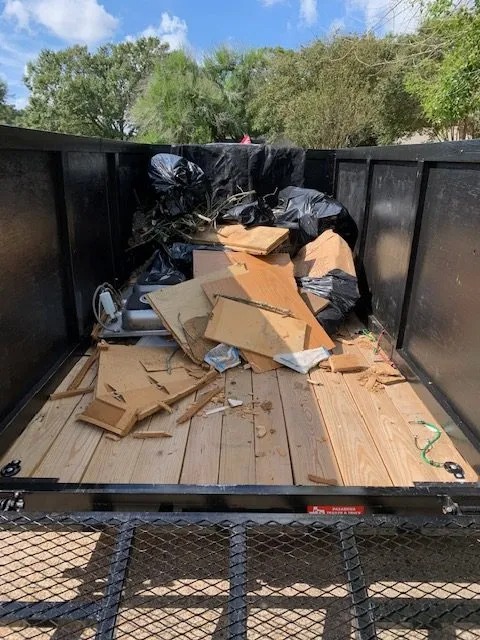 Pełna przyczepa gruzu i śmieci, przygotowana do wywozu. Widoczne deski, worki na śmieci i inne odpady. Metalowa konstrukcja przyczepy i siatka zabezpieczająca ładunek.