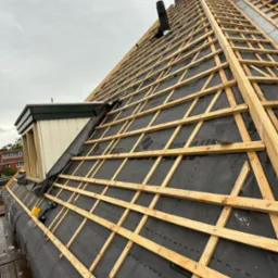 W trakcie montażu drewnianej więźby dachowej na czarnej membranie, widoczny fragment komina i okno dachowe. Na dachu leży wkrętarka.