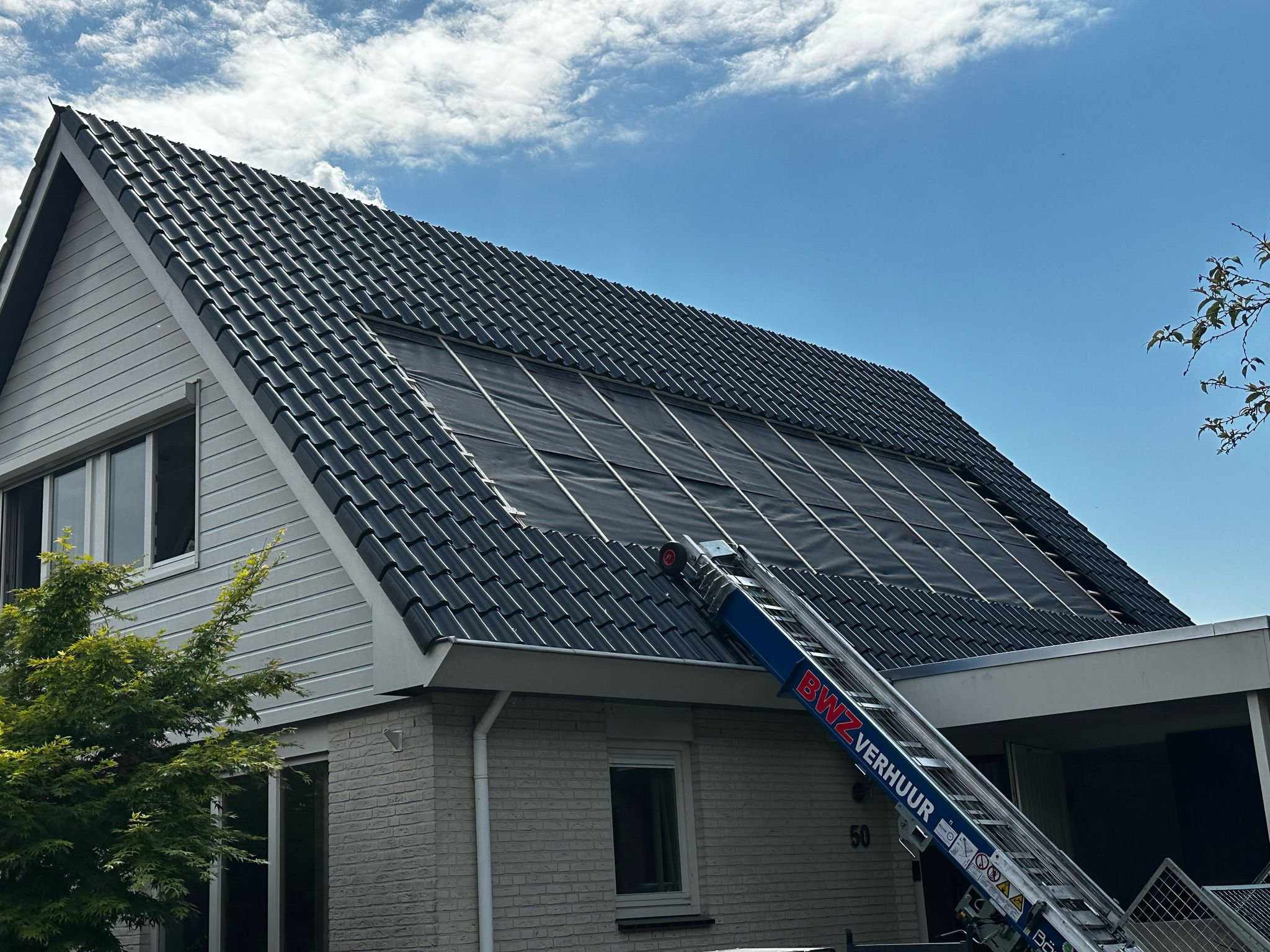 Dom z czarną dachówką, na dachu rozłożona czarna folia. Do dachu przystawiona niebieska drabina z napisem BWZ Verhuur. Błękitne niebo z chmurami w tle.