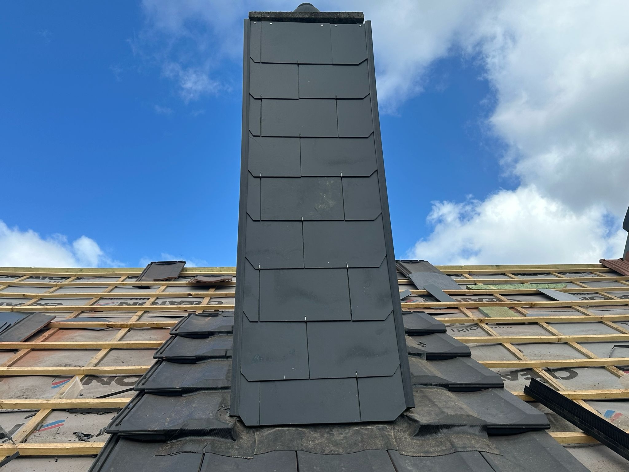 Montaż ciemnej dachówki wokół komina na tle błękitnego nieba. Widoczne elementy konstrukcji dachu i membrany. Dachówka ułożona w regularny wzór.