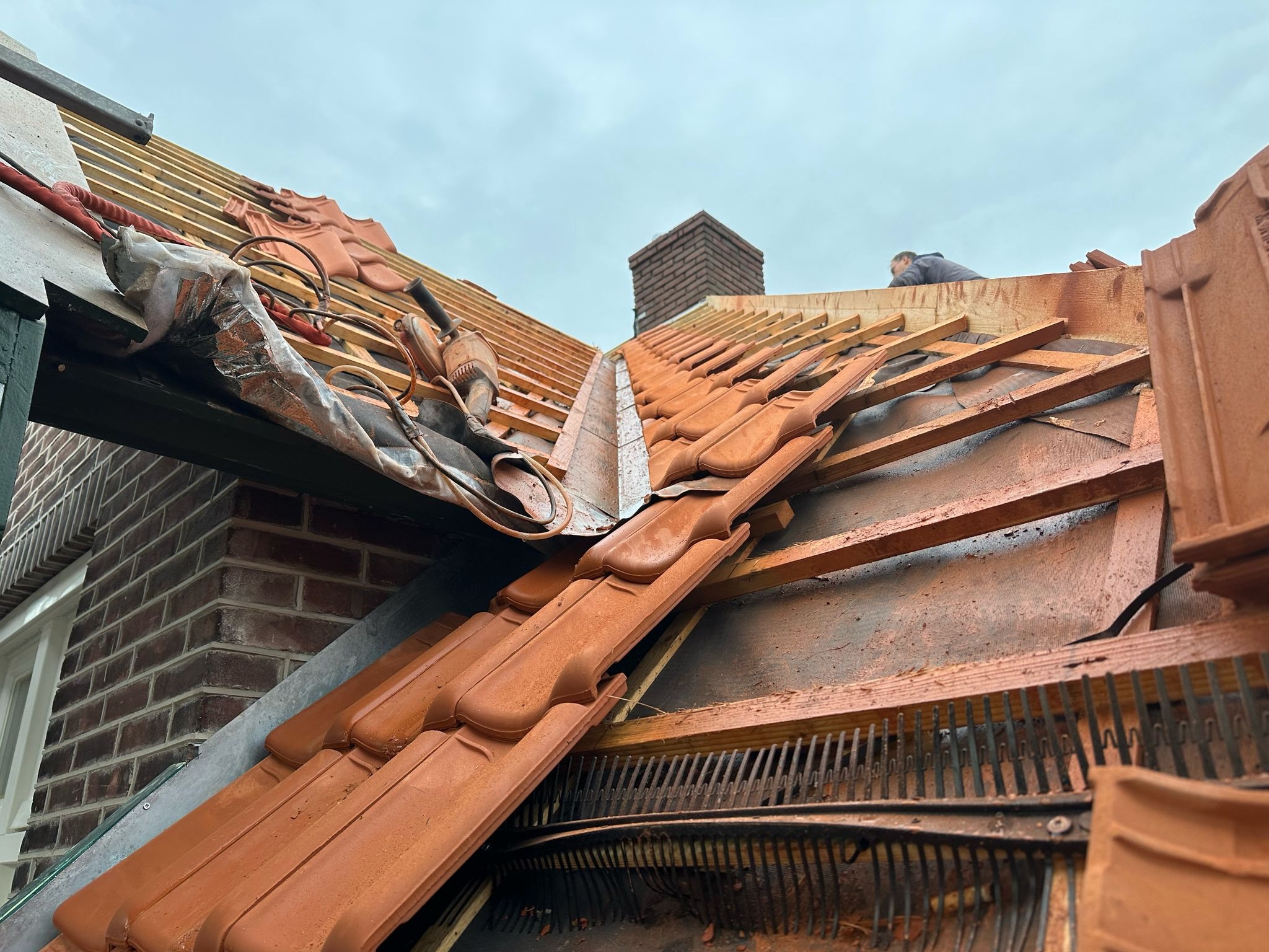Częściowy widok dachu w trakcie krycia czerwoną dachówką. Widoczne elementy więźby, komin w tle i narzędzia dekarskie.