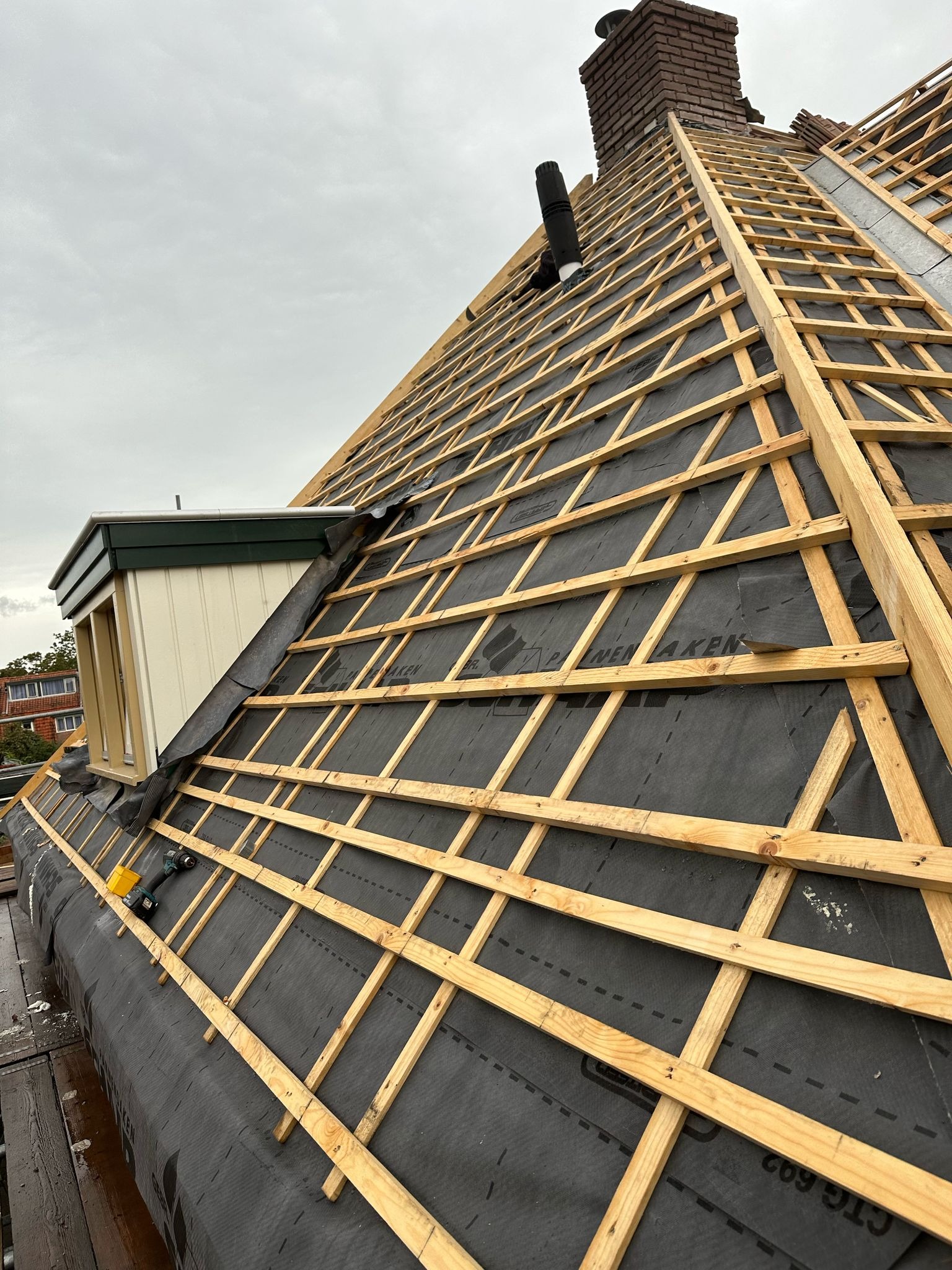 W trakcie montażu drewnianej więźby dachowej na czarnej membranie, widoczny fragment komina i okno dachowe. Na dachu leży wkrętarka.