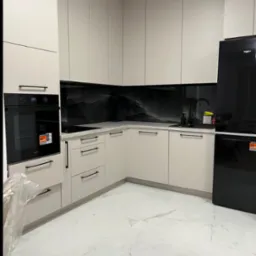 Kremowa kuchnia na wymiar z czarnym AGD i marmurową podłogą. Minimalistyczne uchwyty i panel nad blatem w stylu marmuru. Nowoczesne wykończenie.