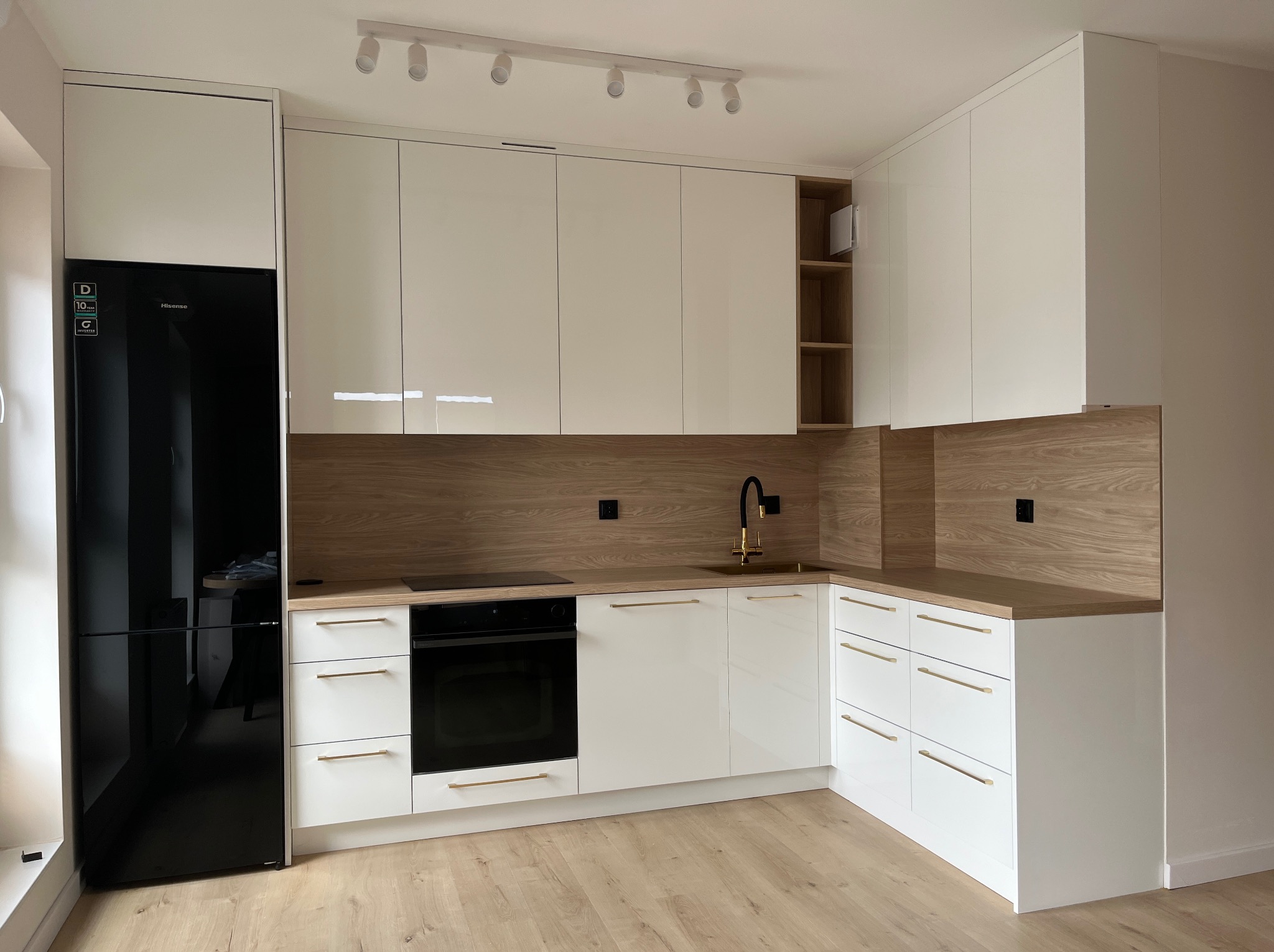 Nowoczesna kuchnia na wymiar z białymi, lakierowanymi frontami, drewnianym blatem i czarną lodówką. Złote uchwyty i armatura dodają elegancji.