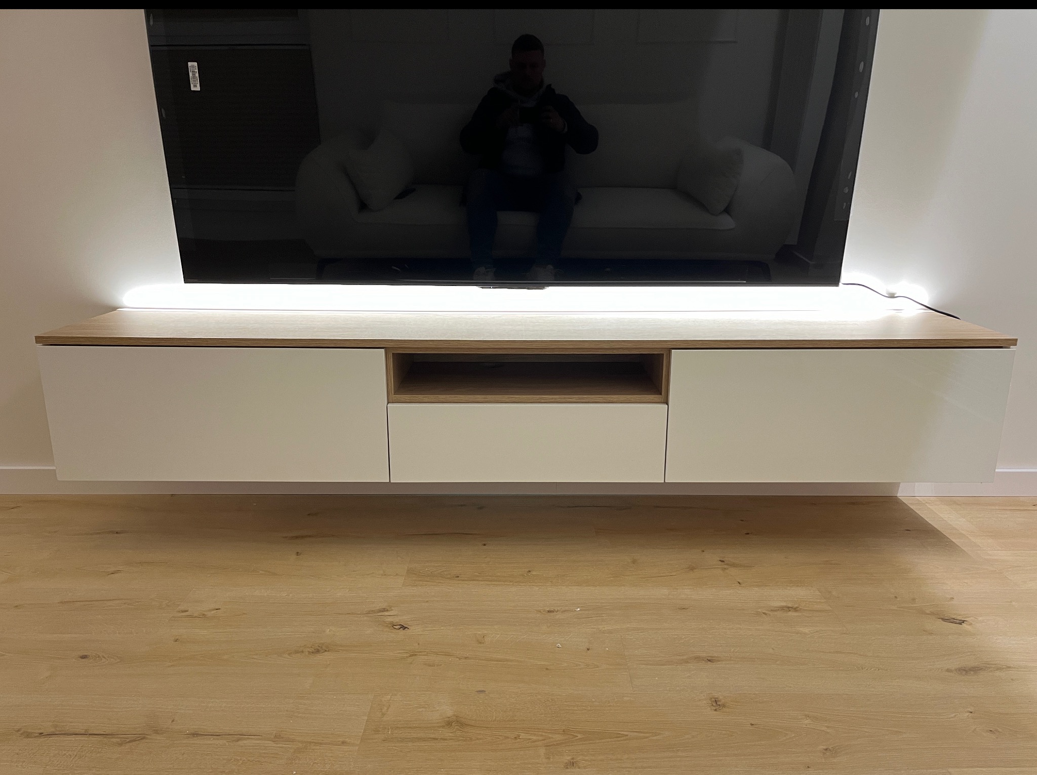 Nowoczesna szafka RTV wisząca, z oświetleniem LED, pod telewizorem. Jasne drewno i białe fronty. Parkiet na podłodze. Widoczne odbicie osoby w ekranie TV.