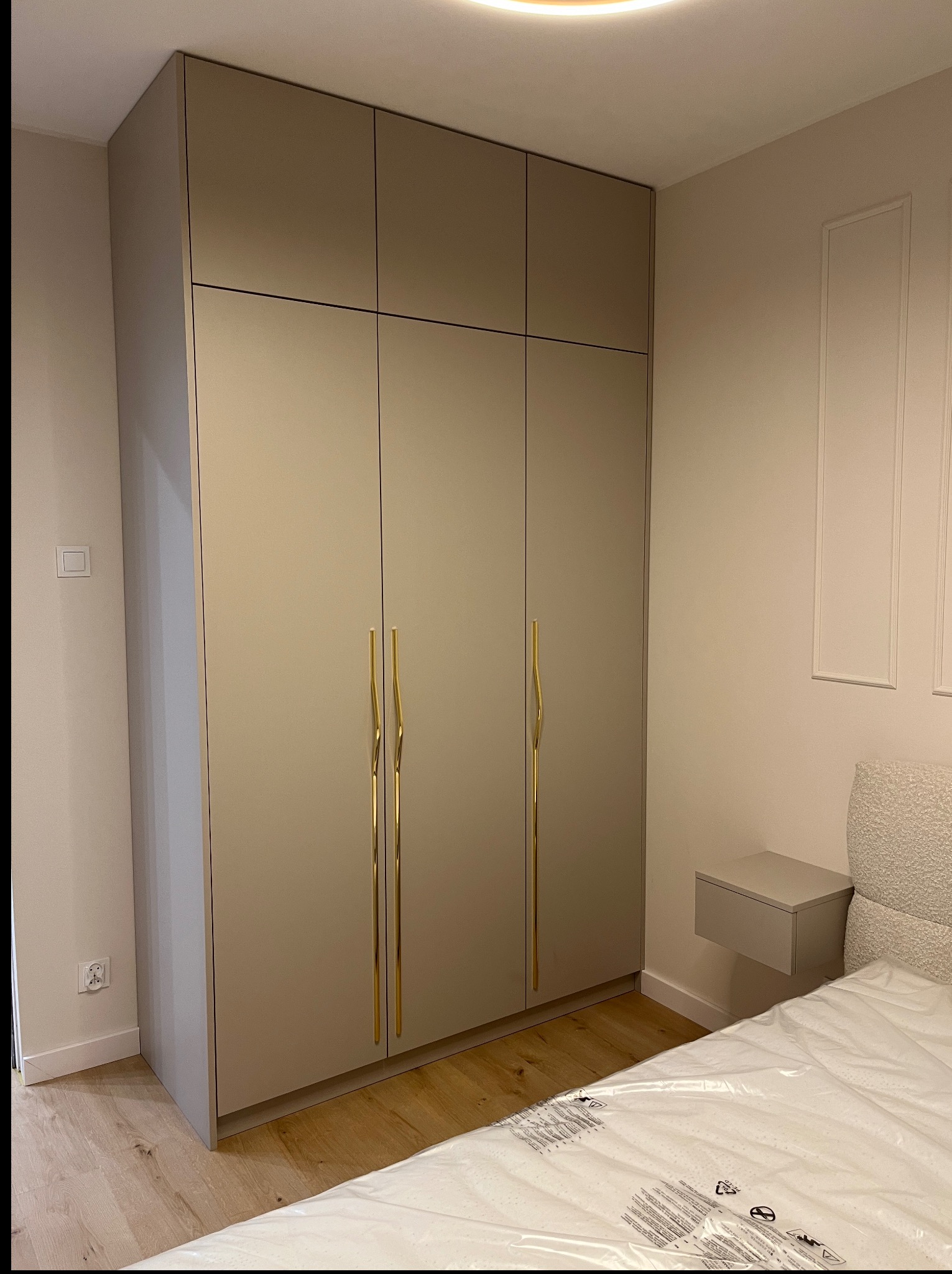 Beżowa szafa na wymiar z złotymi, zakrzywionymi uchwytami w sypialni. Widoczny fragment łóżka z zapakowanym materacem i szafka nocna w kolorze szafy.