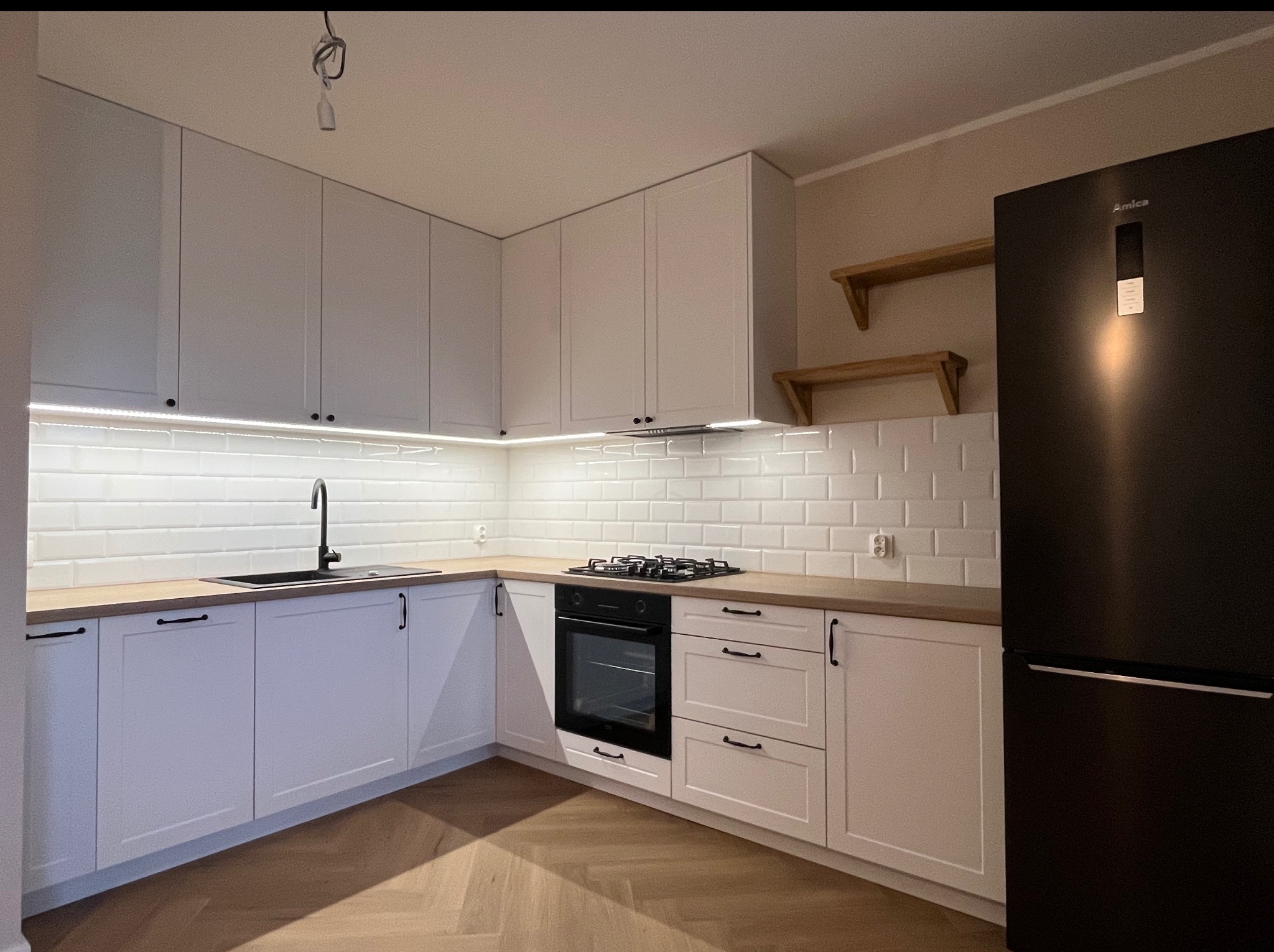 Jasna kuchnia z białymi frontami, drewnianym blatem i czarnymi akcentami: zlew, bateria, piekarnik, lodówka. Na ścianie białe płytki imitujące cegłę.