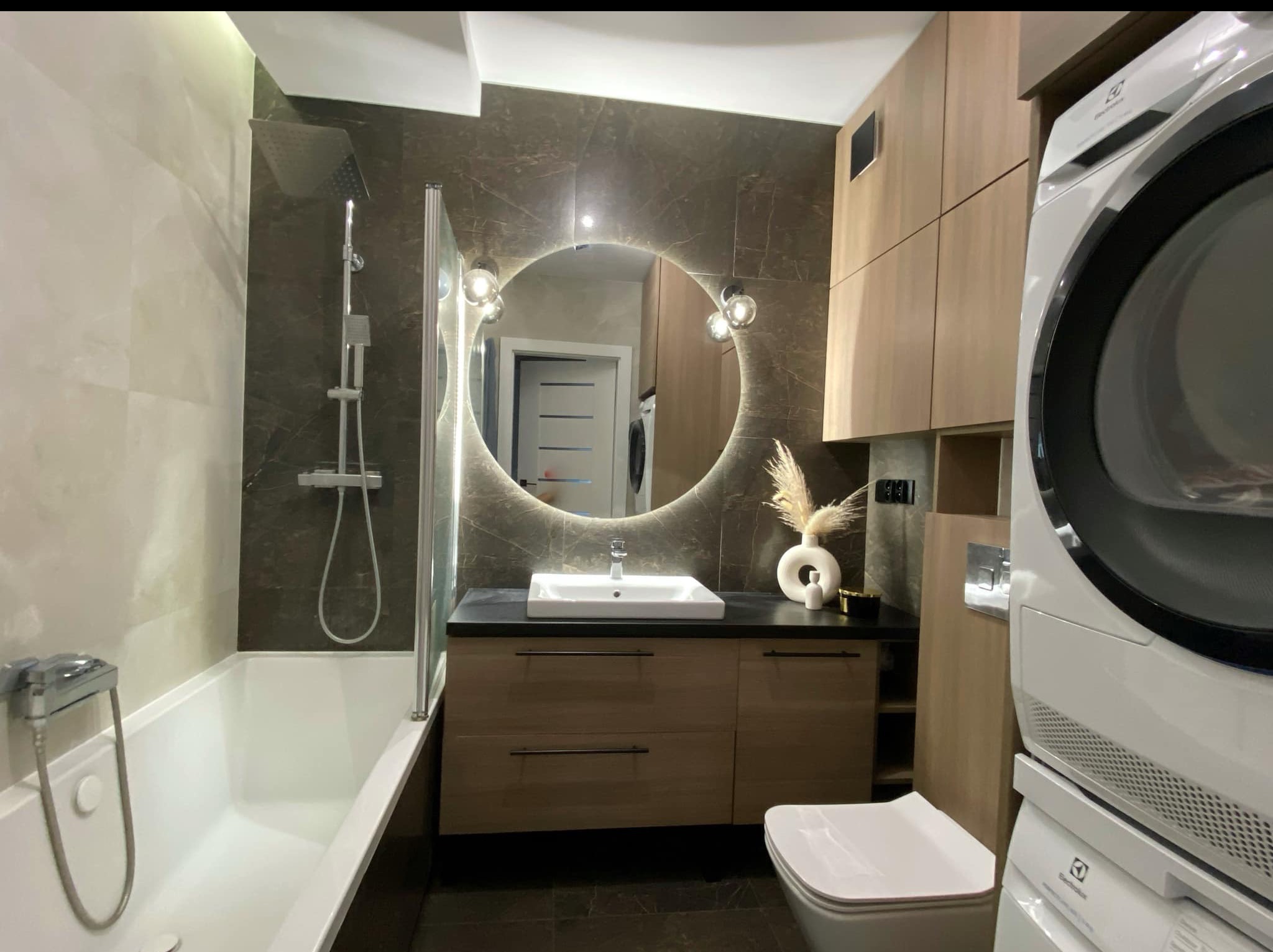 Nowoczesna łazienka w Wasilkowie z okrągłym lustrem LED, drewnianymi szafkami, białą armaturą i pralko-suszarką. Eleganckie wykończenie marmurową płytką.