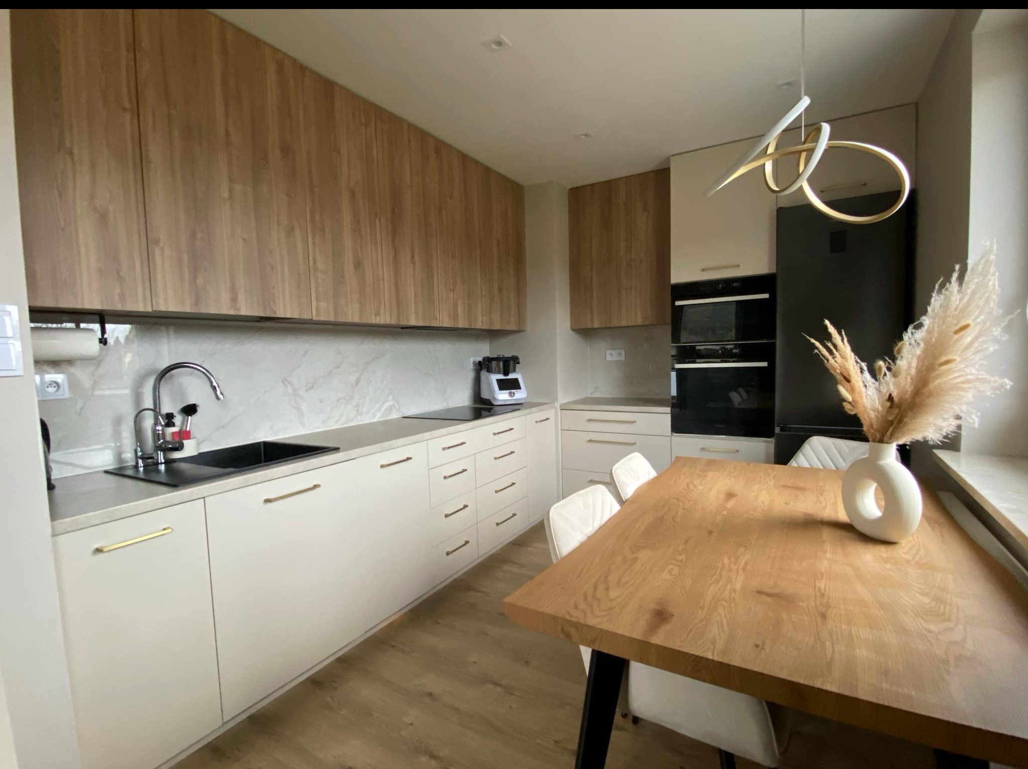 Jasna kuchnia z drewnianymi akcentami: beżowe szafki dolne z złotymi uchwytami, górne szafki w odcieniu drewna, stół i dekoracyjny wazon na blacie.