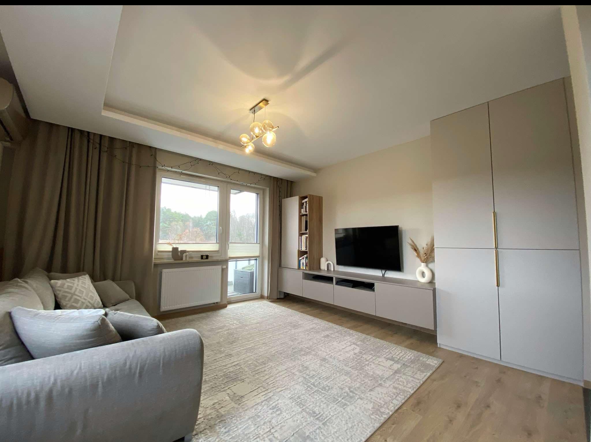 Jasny salon z szarą sofą, dywanem i meblami na wymiar w odcieniach beżu. Widoczne okno i lampa sufitowa. Minimalistyczny design z elementami dekoracyjnymi.