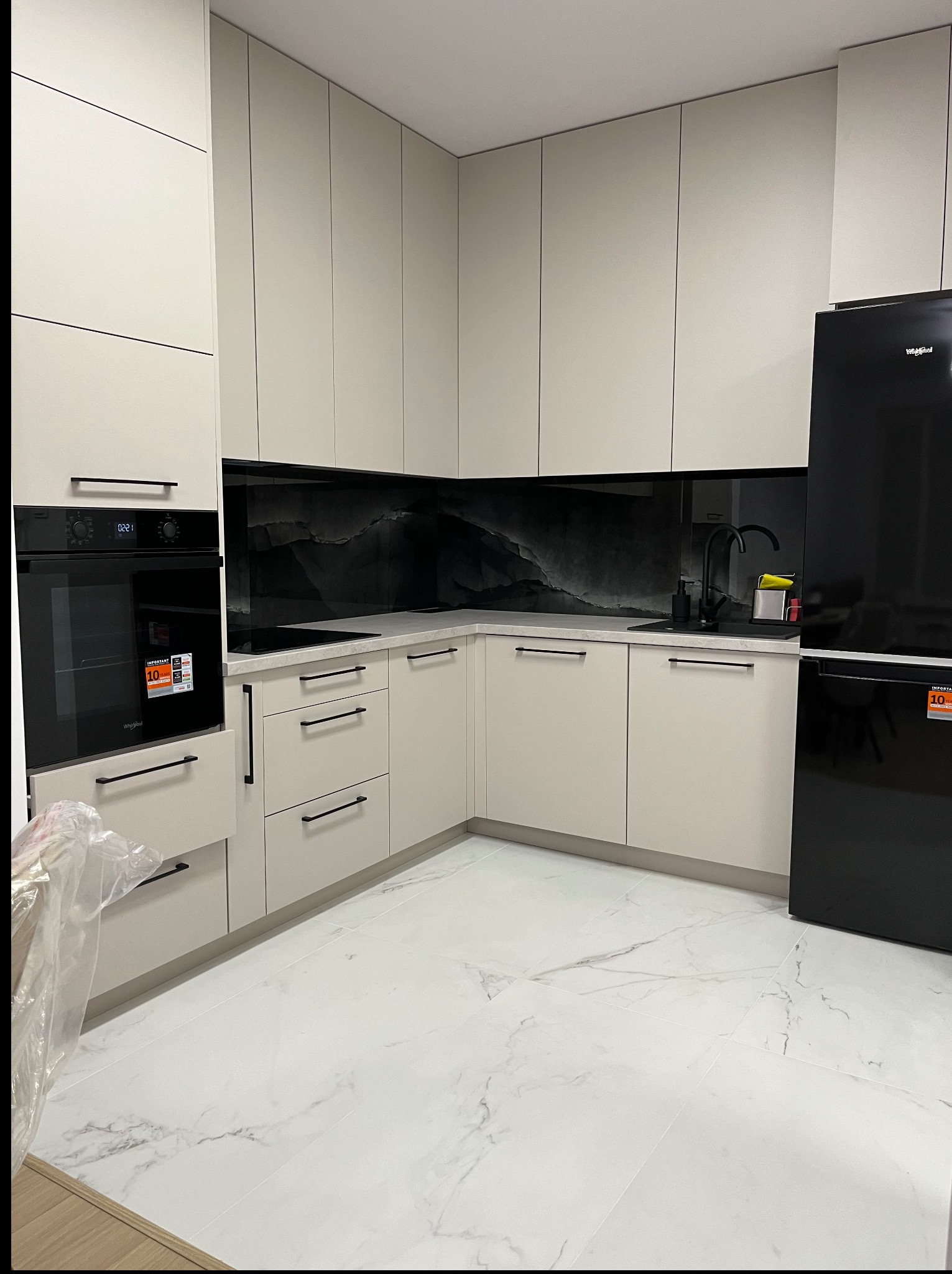 Kremowa kuchnia na wymiar z czarnym AGD i marmurową podłogą. Minimalistyczne uchwyty i panel nad blatem w stylu marmuru. Nowoczesne wykończenie.