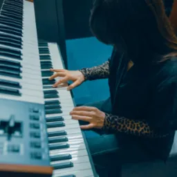 Kobieta gra na klawiszach elektronicznych w studio nagrań; widoczne dłonie na klawiaturze, panel sterowania po lewej stronie. Ubrana w bluzkę w panterkę.