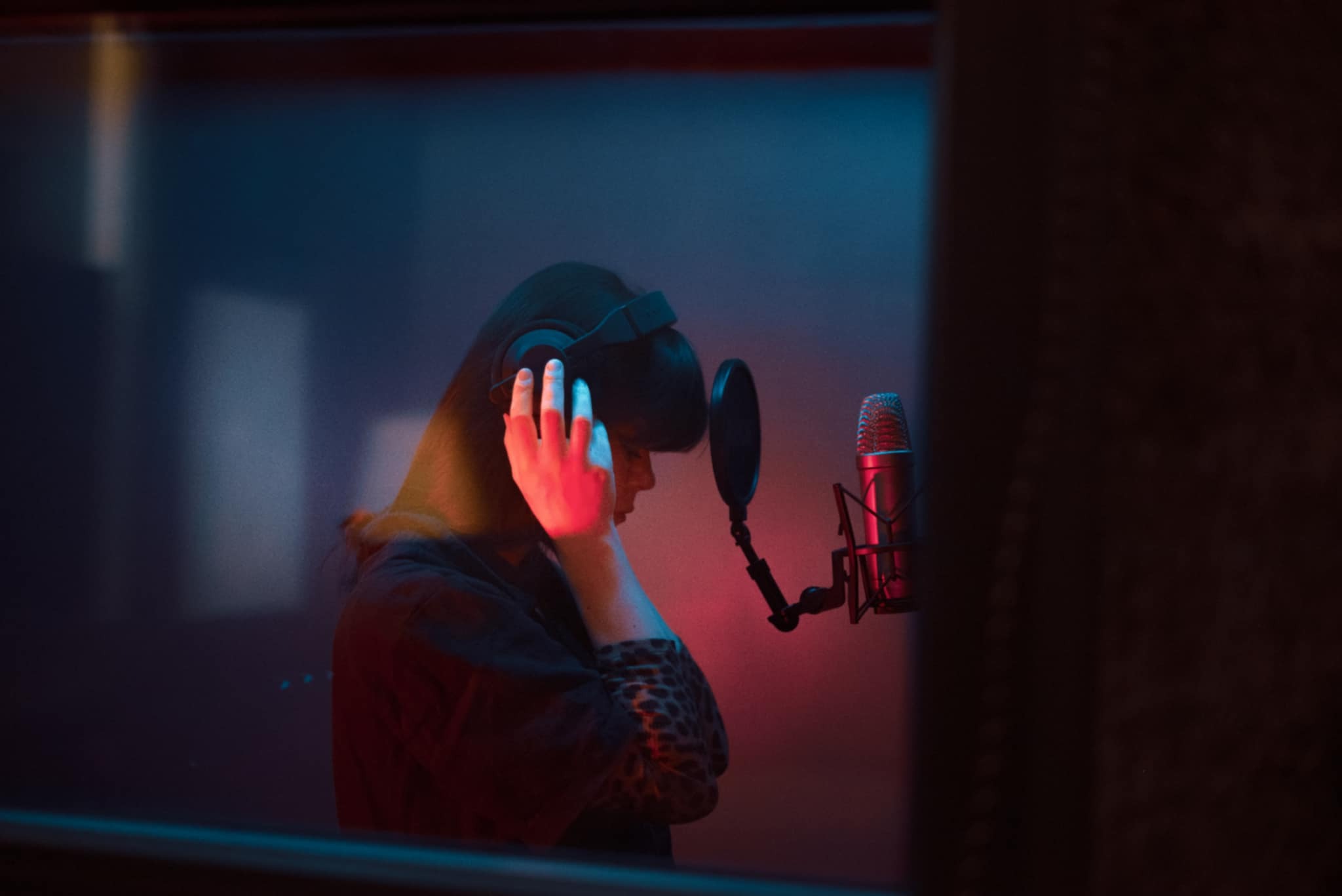 Kobieta w słuchawkach, przed mikrofonem w kabinie studia nagrań, oświetlonej czerwono-niebieskim światłem. Widok zza szyby.