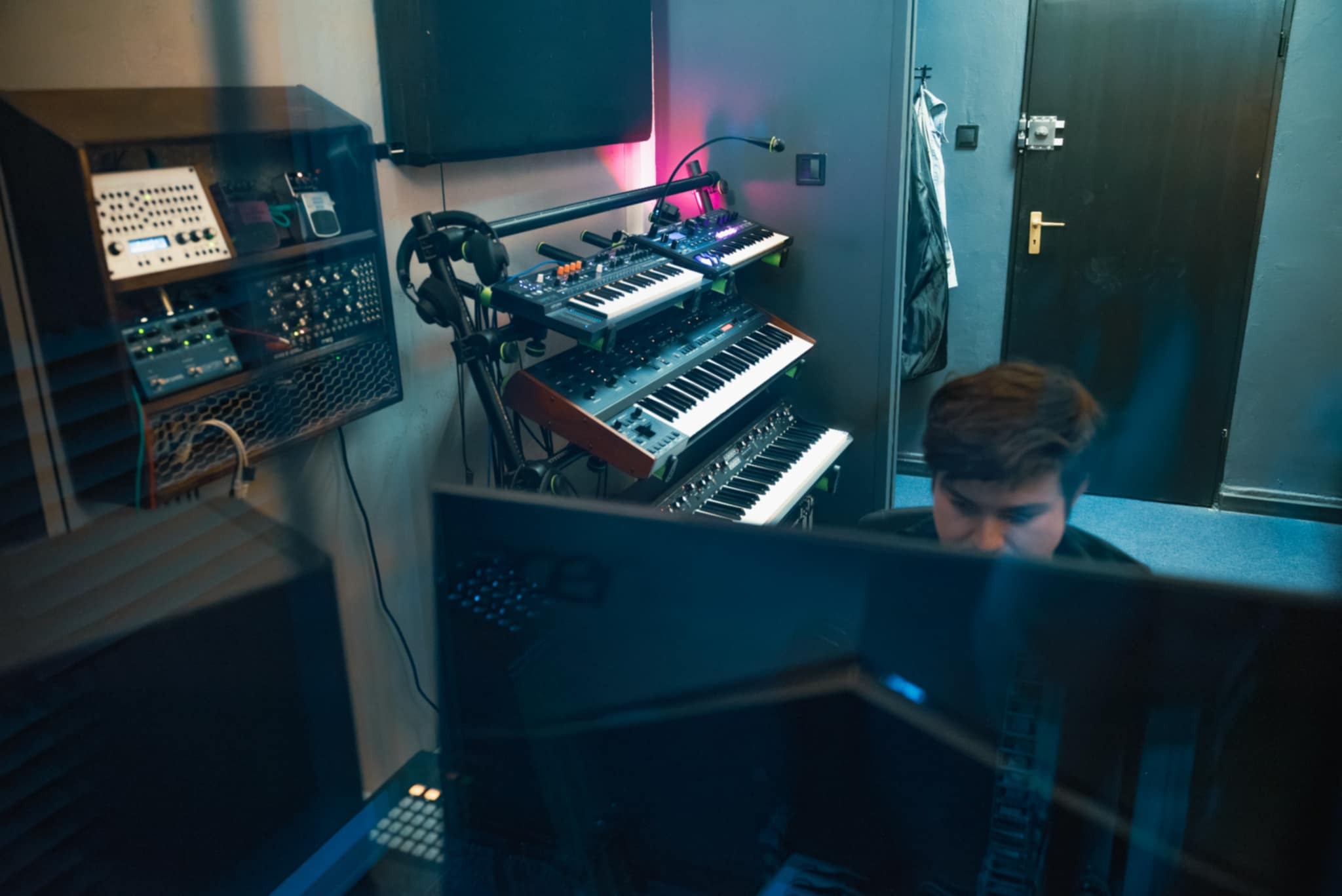 Wnętrze studia nagrań z syntezatorami, rackiem sprzętowym i monitorem. Widoczny fragment osoby pracującej przy stanowisku. Ściany w odcieniach szarości i niebieskiego.