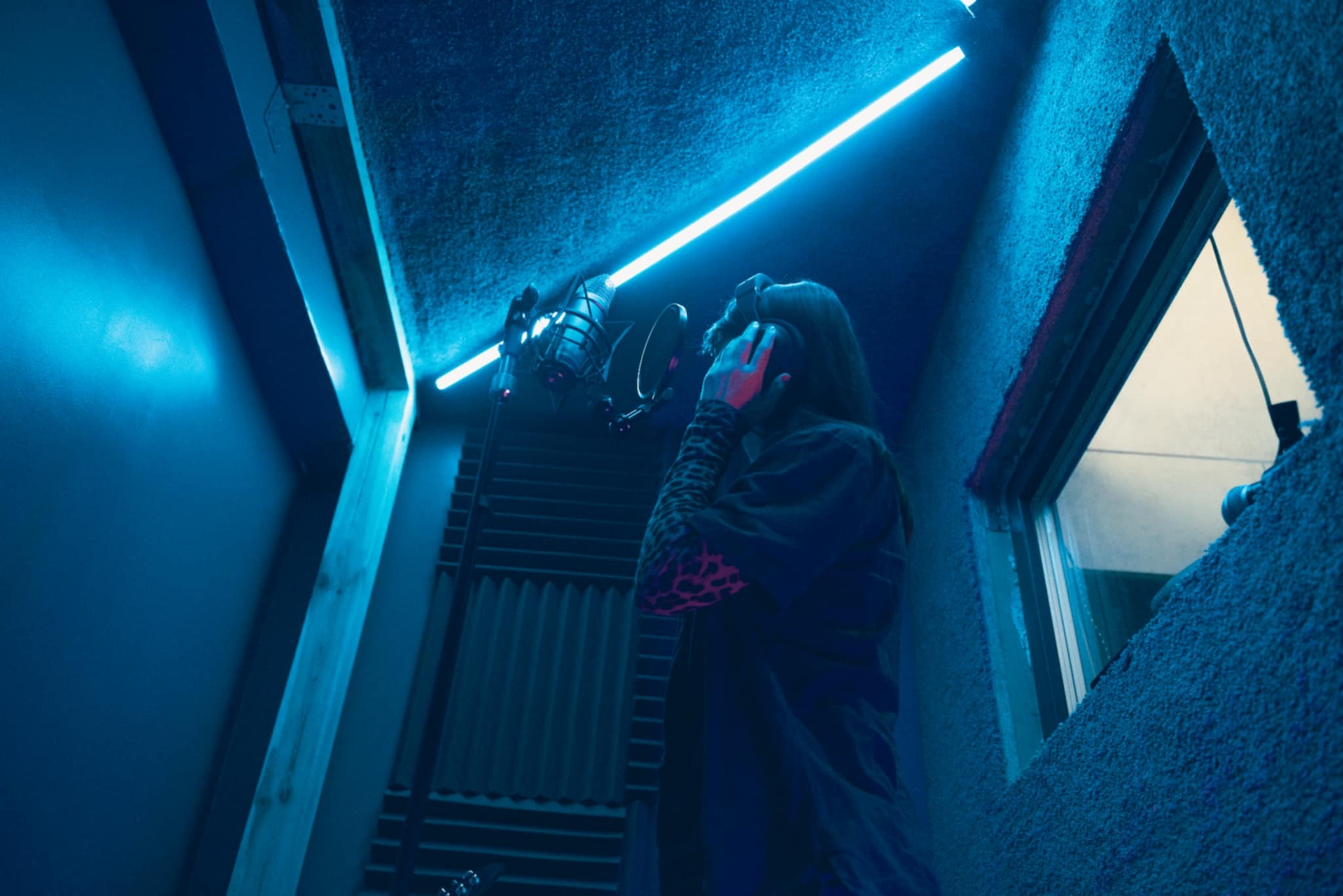 Osoba w słuchawkach przed mikrofonem w niebieskim oświetleniu studia nagrań. Widoczne panele akustyczne i okno odsłuchowe.