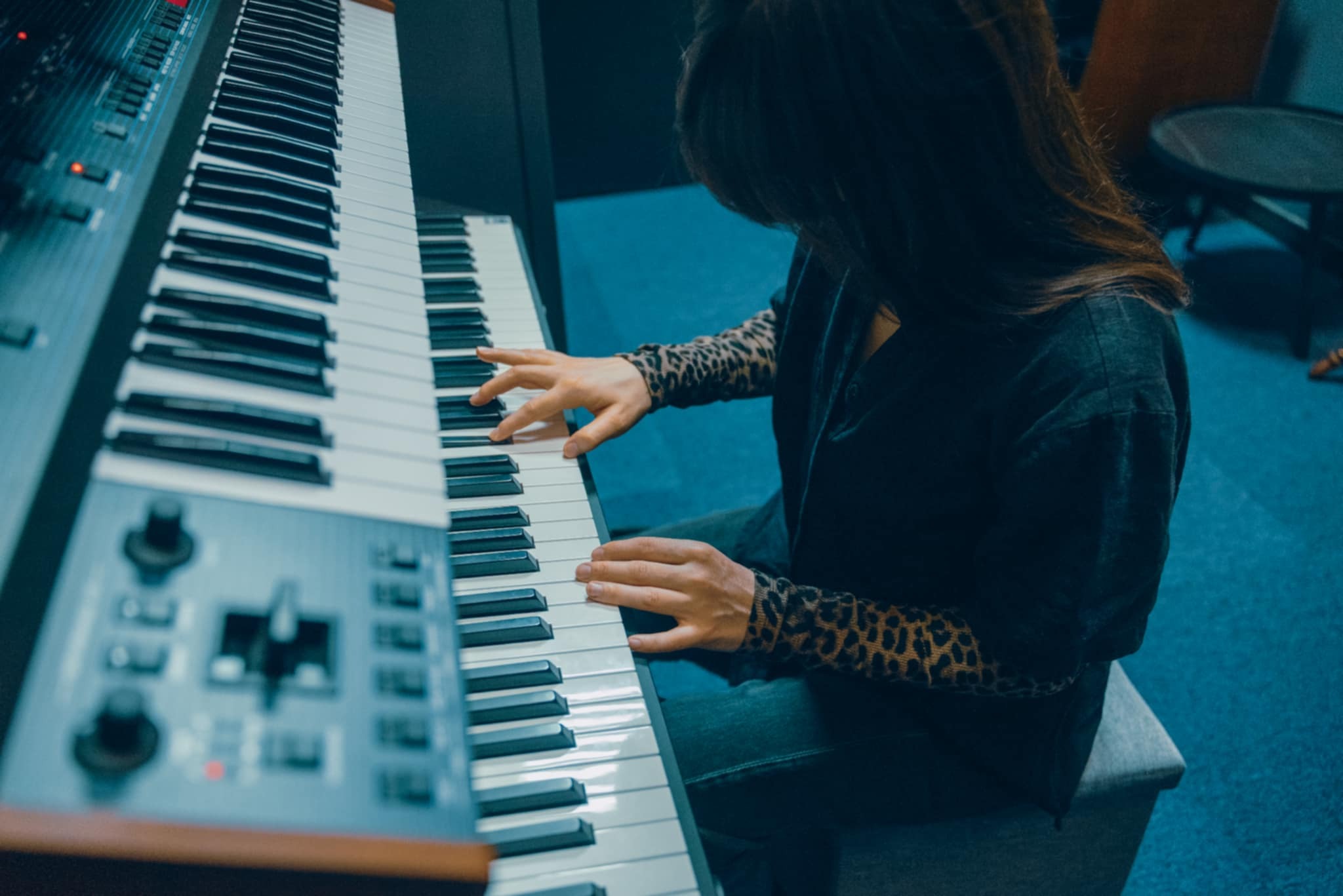 Kobieta gra na klawiszach elektronicznych w studio nagrań; widoczne dłonie na klawiaturze, panel sterowania po lewej stronie. Ubrana w bluzkę w panterkę.