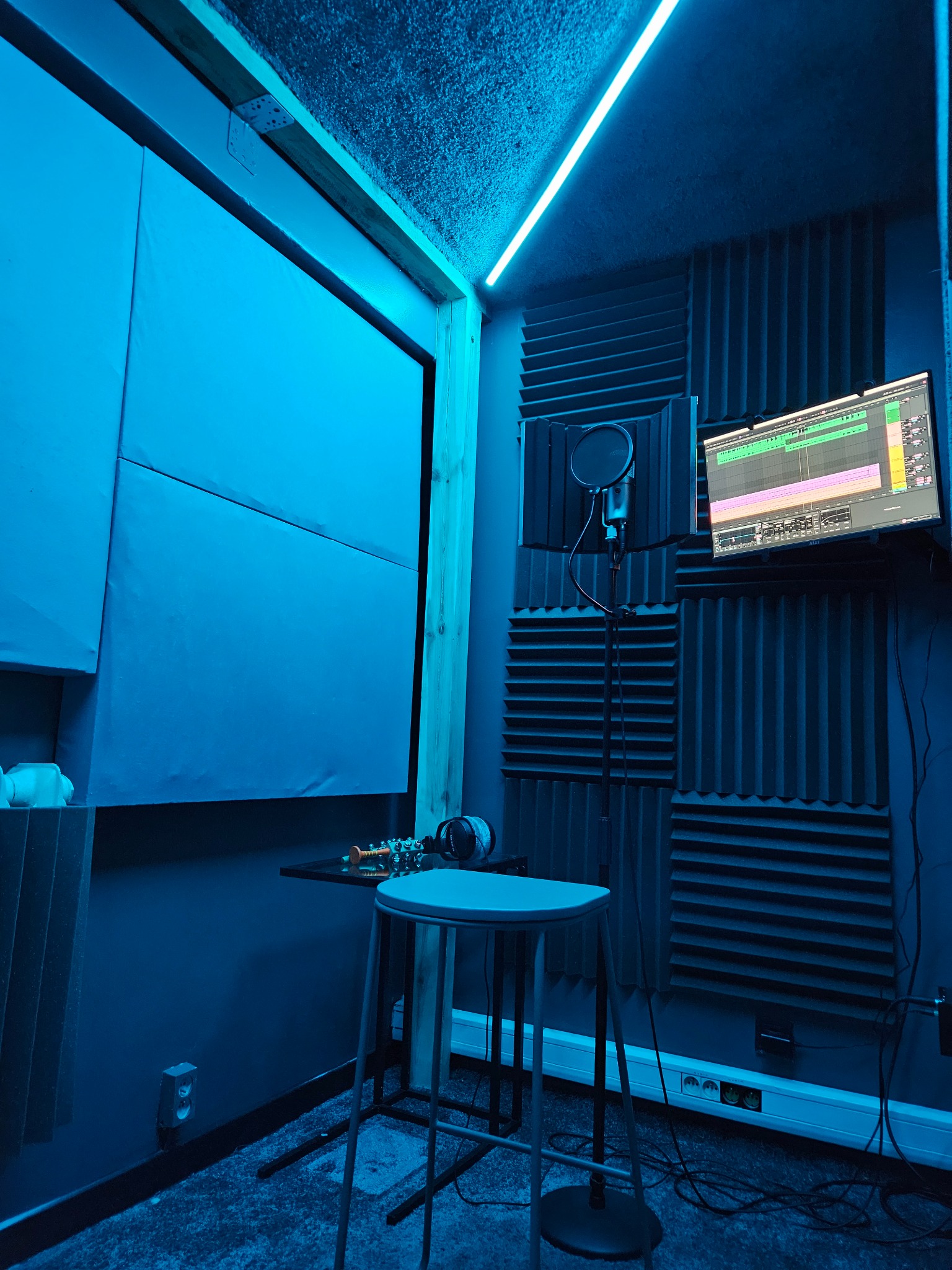 Wnętrze studia nagrań w niebieskiej tonacji: mikrofon z pop filtrem, monitor z oprogramowaniem DAW, panele akustyczne i ledowe oświetlenie.