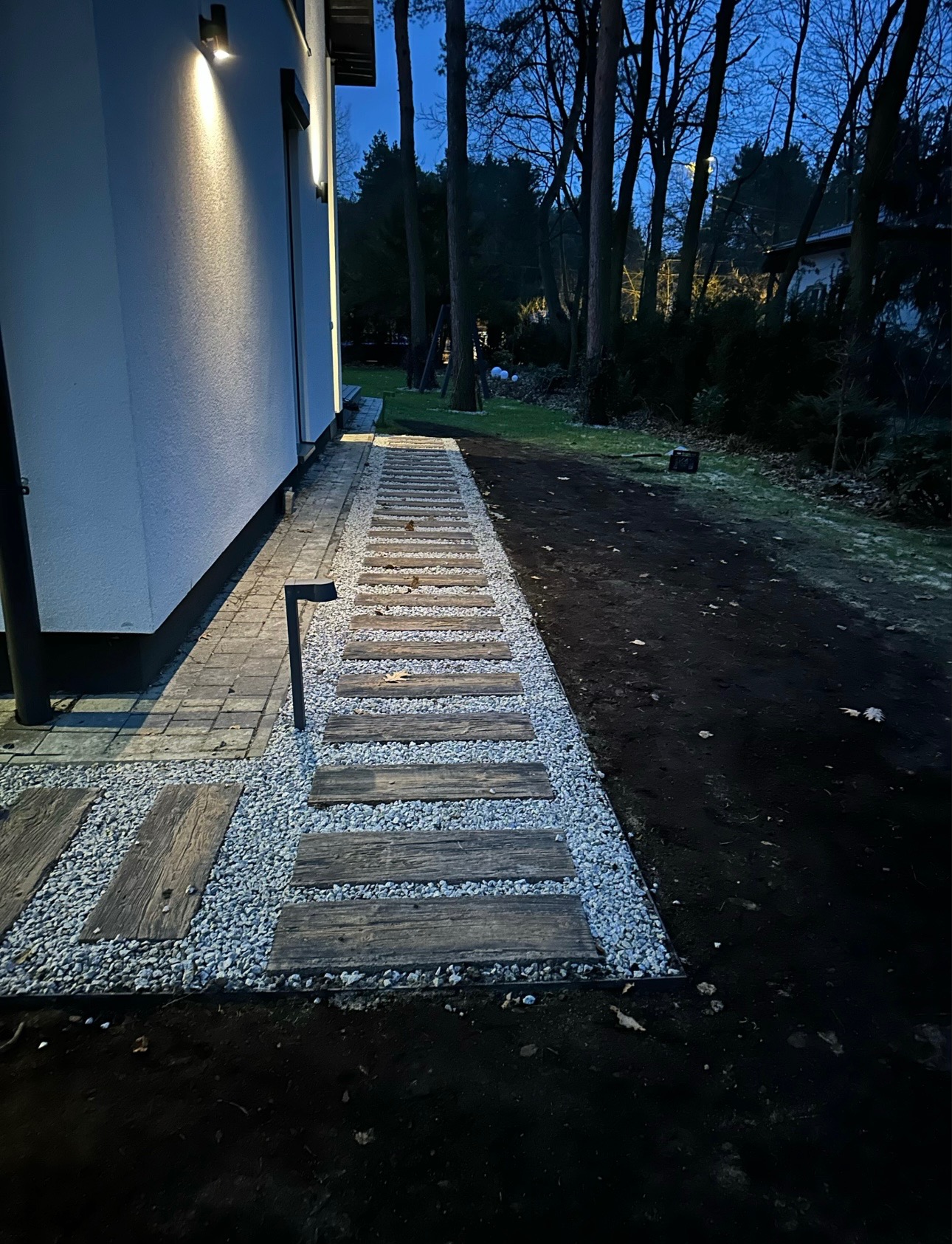 Ścieżka z drewnianych płyt i grysu przy domu, oświetlona lampami. Nowoczesny ogród w wieczornym świetle, widok na drzewa w tle.